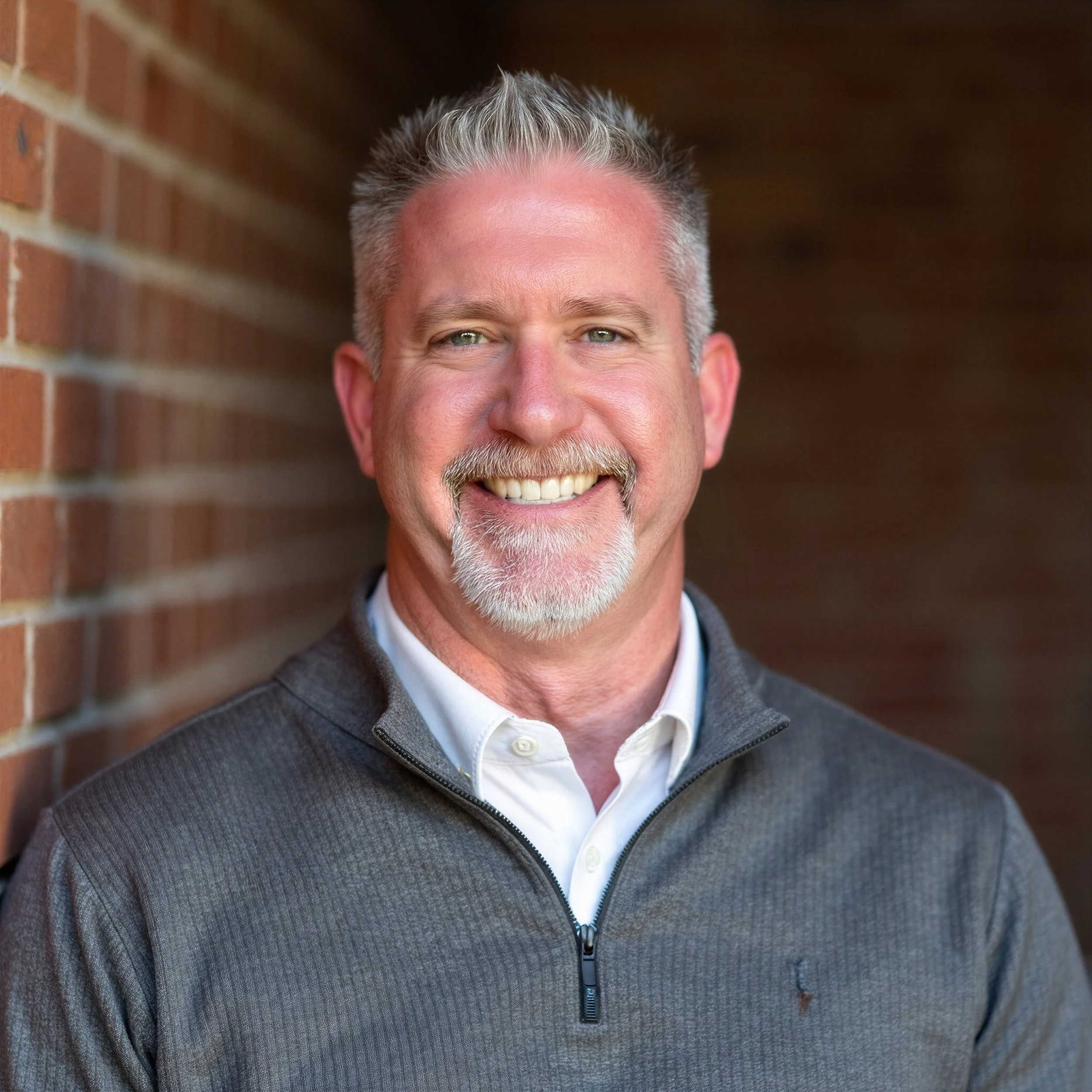 Close-up of a man's face showing his eyes, smiling mouth, and gray hair with a beard, standing against a brick wall.