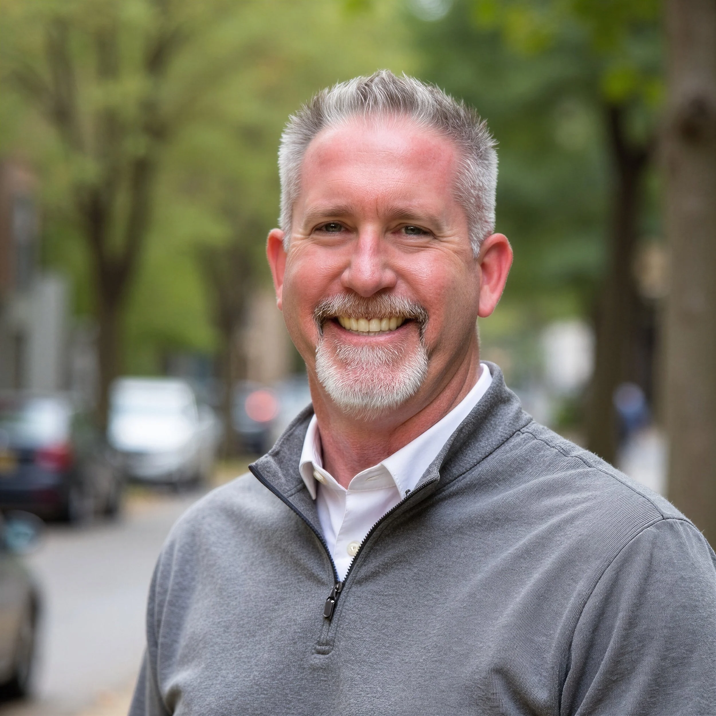 A middle-aged man with short gray hair and a goatee smiling outdoors. He is wearing a gray zip-up sweater over a white collared shirt. The background is blurred with green trees and some cars visible.