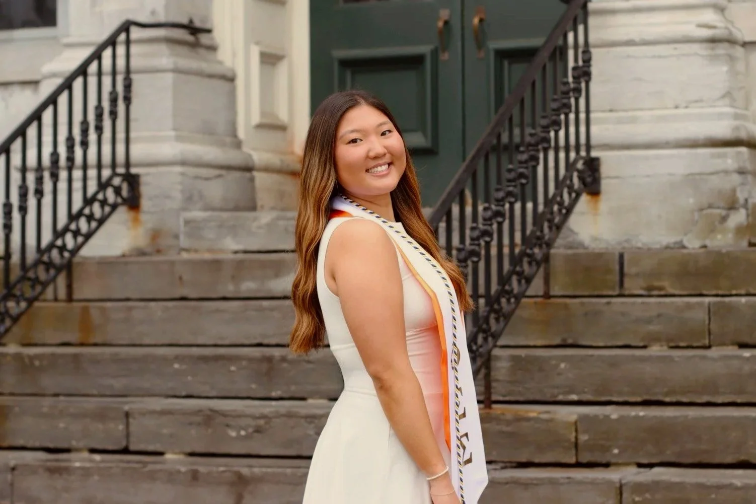 Young woman in a white dress with a graduation stole, standing on steps outside a historic building, smiling at the camera.