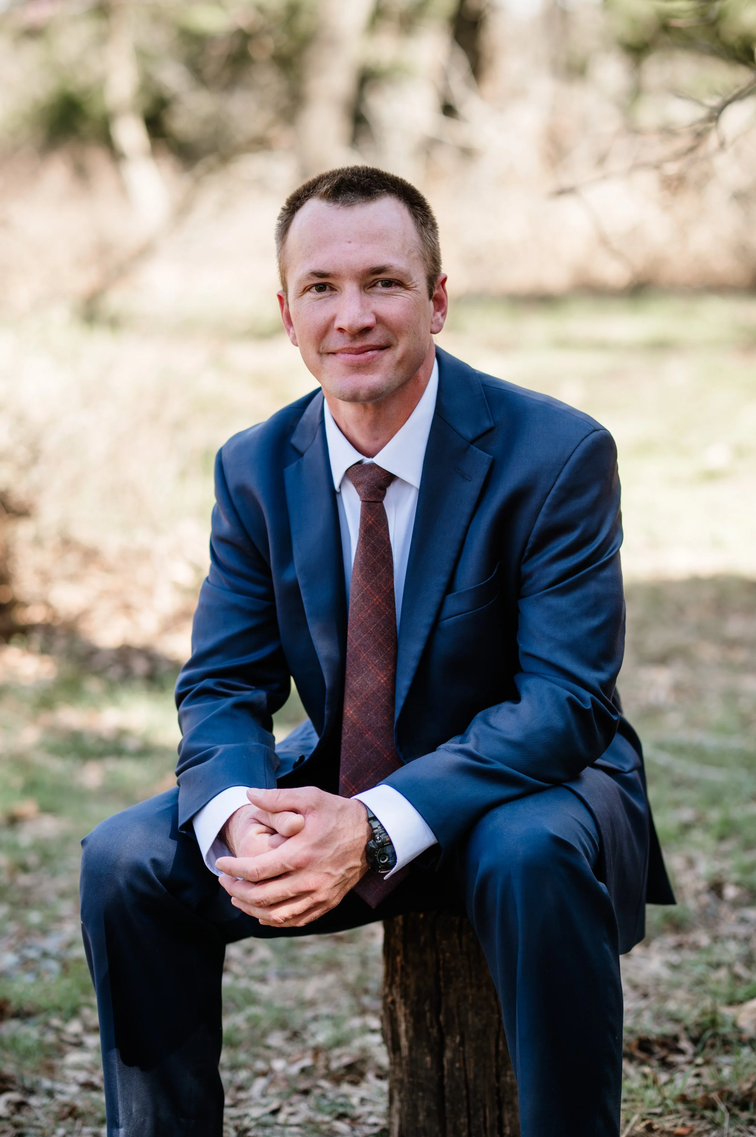 A man in a navy blue suit, white shirt, and brown tie sitting on a tree stump outdoors with a background of trees and grass, smiling at the camera.