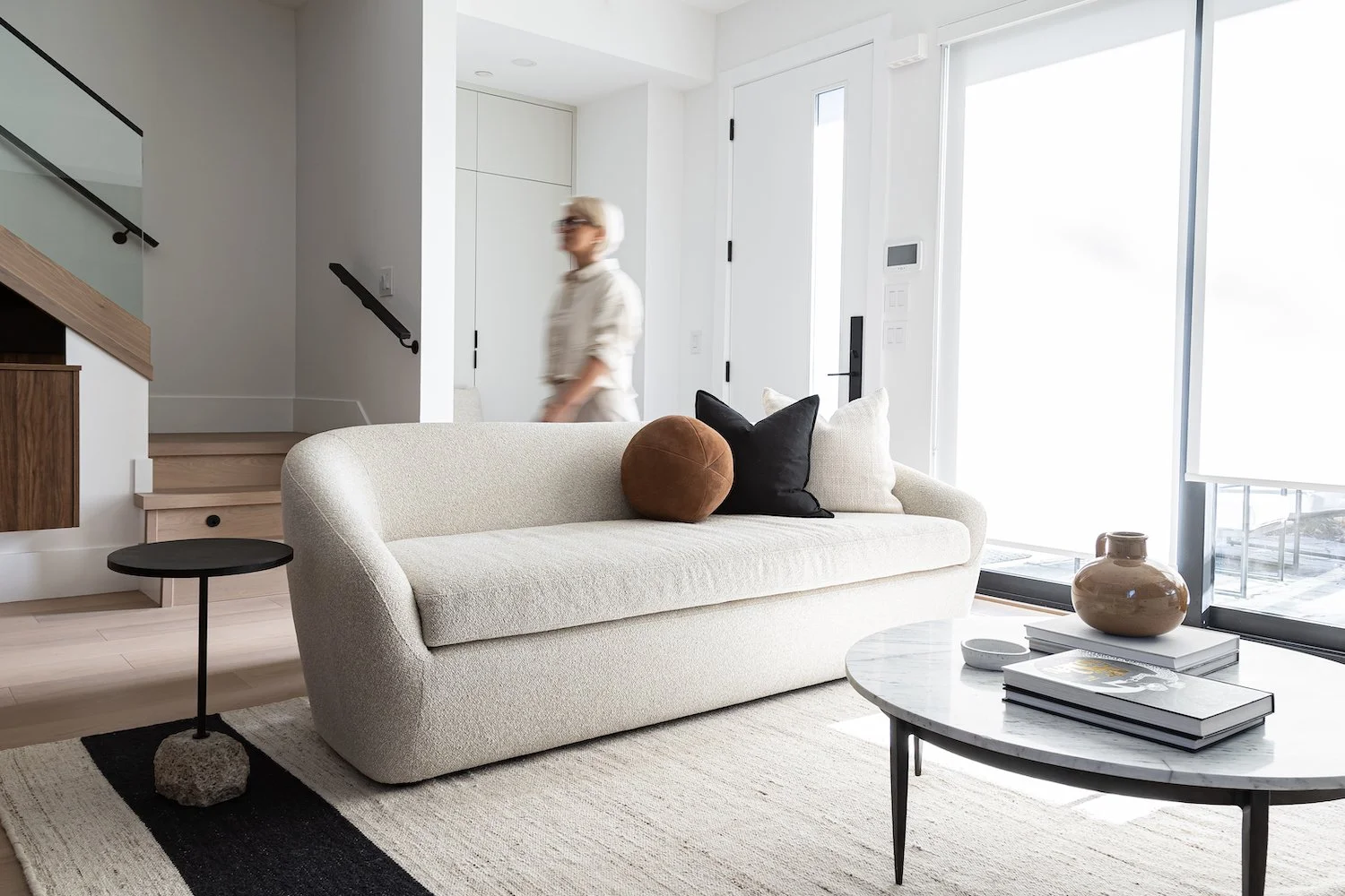 Modern living room with a beige sofa adorned with black, brown, and white pillows, a round marble coffee table with books and a vase, and a large window letting in bright light.