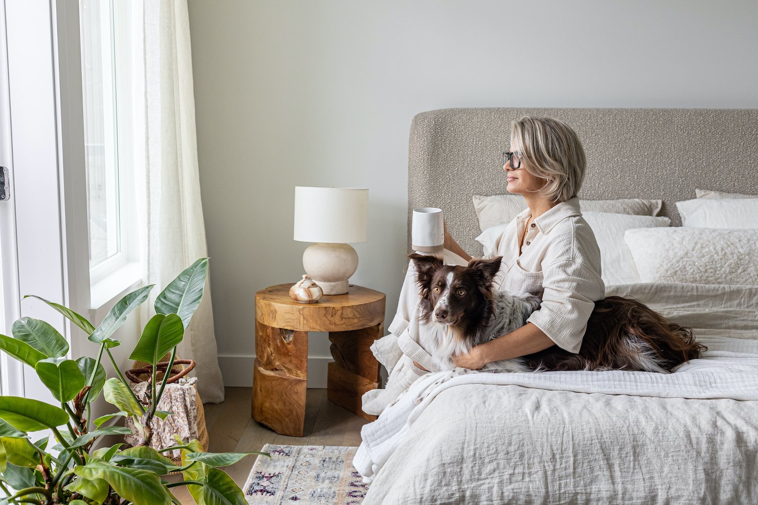 Beautifully staged primary bedroom with neutral linen bedding, rustic wood nightstand, ceramic table lamp, and lush greenery in a bright, airy space
