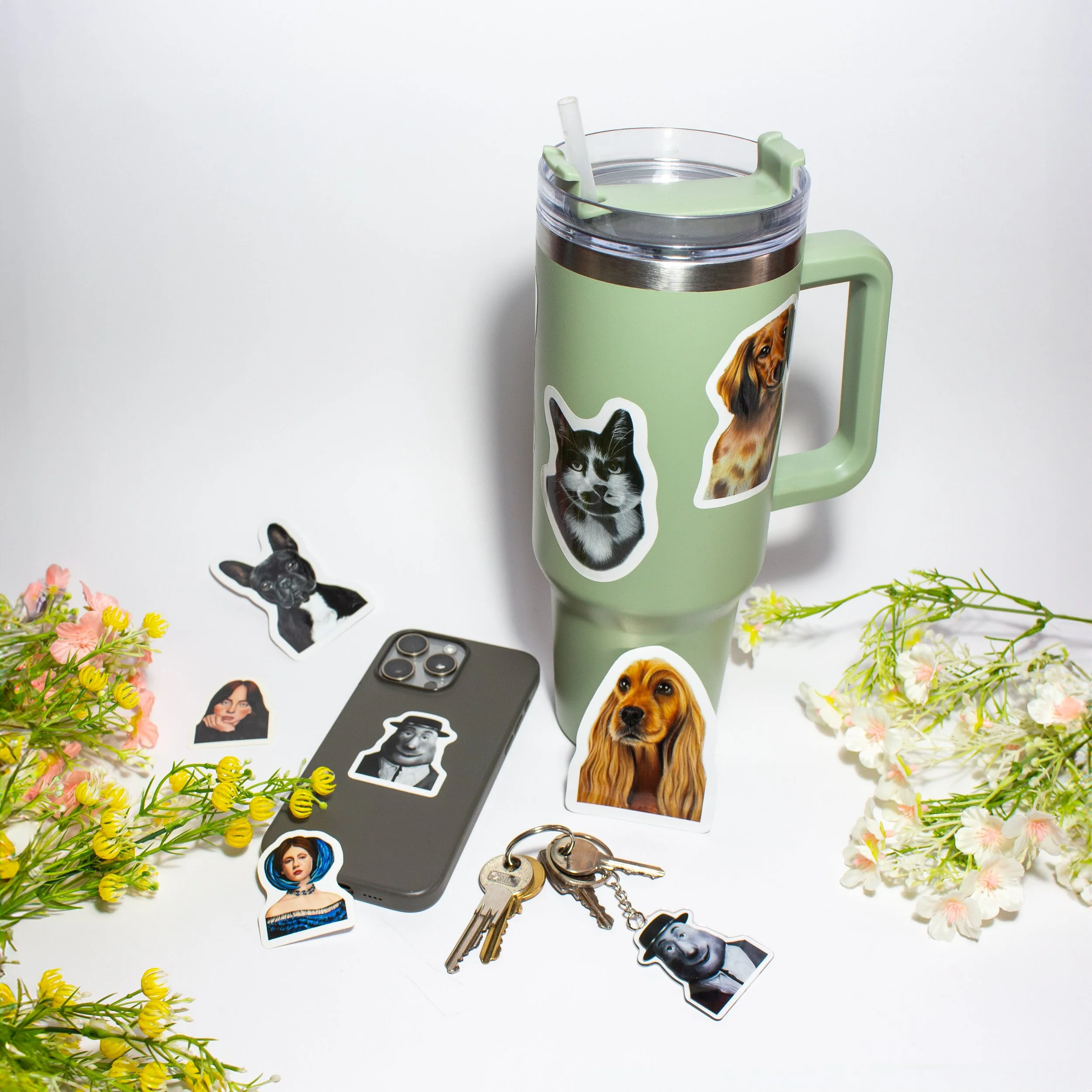 A collection of colorful paper stickers of various people, including a woman with blue hair, a dog, a cat, and a man with a hat, scattered on a white surface alongside a green travel mug with a dog sticker, a smartphone, and a set of keys, surrounded by flowers.