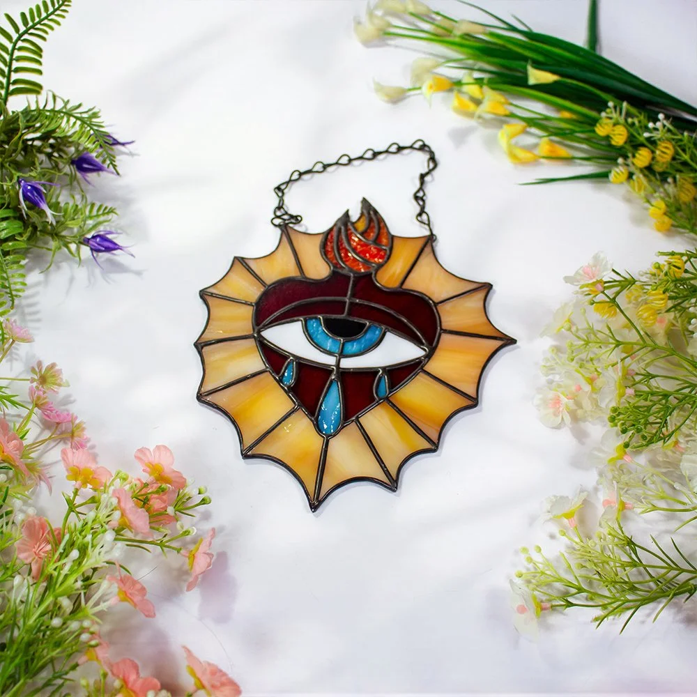 A stained glass ornament featuring a central eye with a tear, surrounded by a fiery flame and a web-like border, is surrounded by flowers and greenery on a white background.