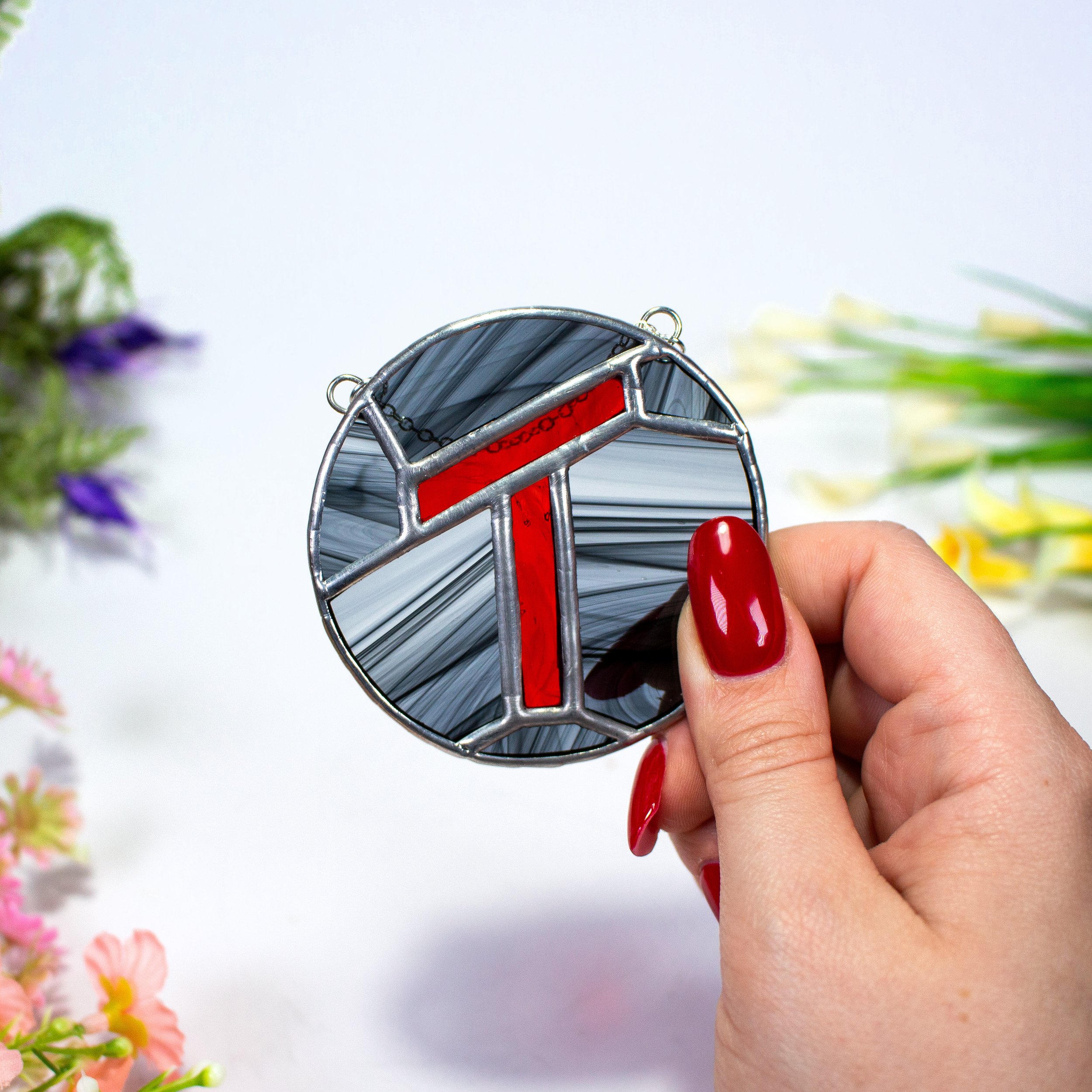Close-up of a person's hand holding a small, metallic, circular object with a red stripe and chain, with blurred flowers in the background.