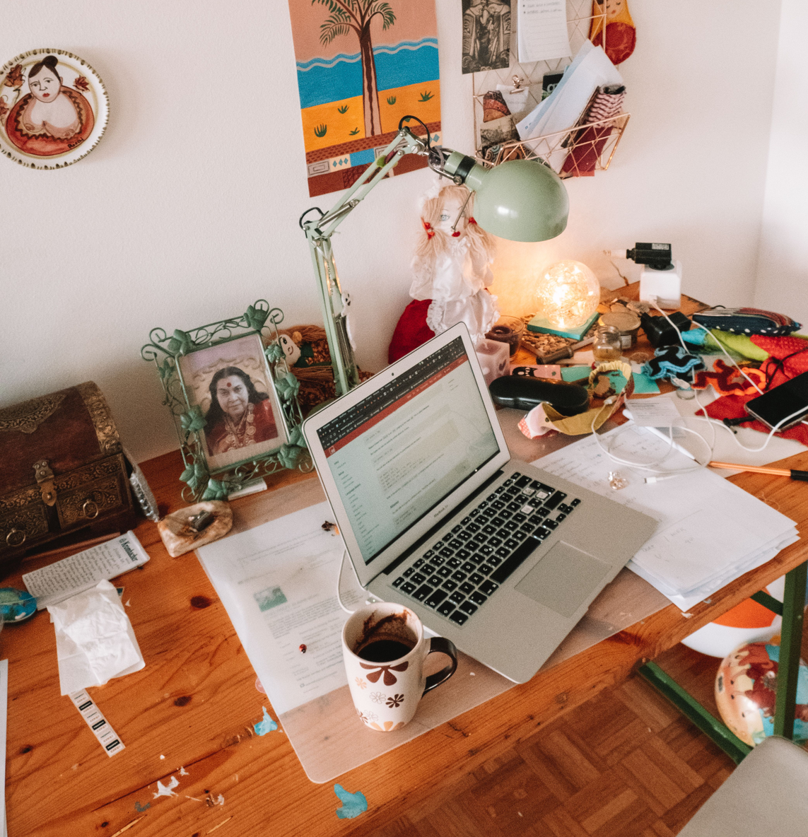 A cluttered wooden desk with a silver laptop, a coffee mug, papers, and various objects including a framed photo and a decorative doll, with a white wall in the background decorated with colorful artwork and a small wire shelf holding documents and a red cloth.
