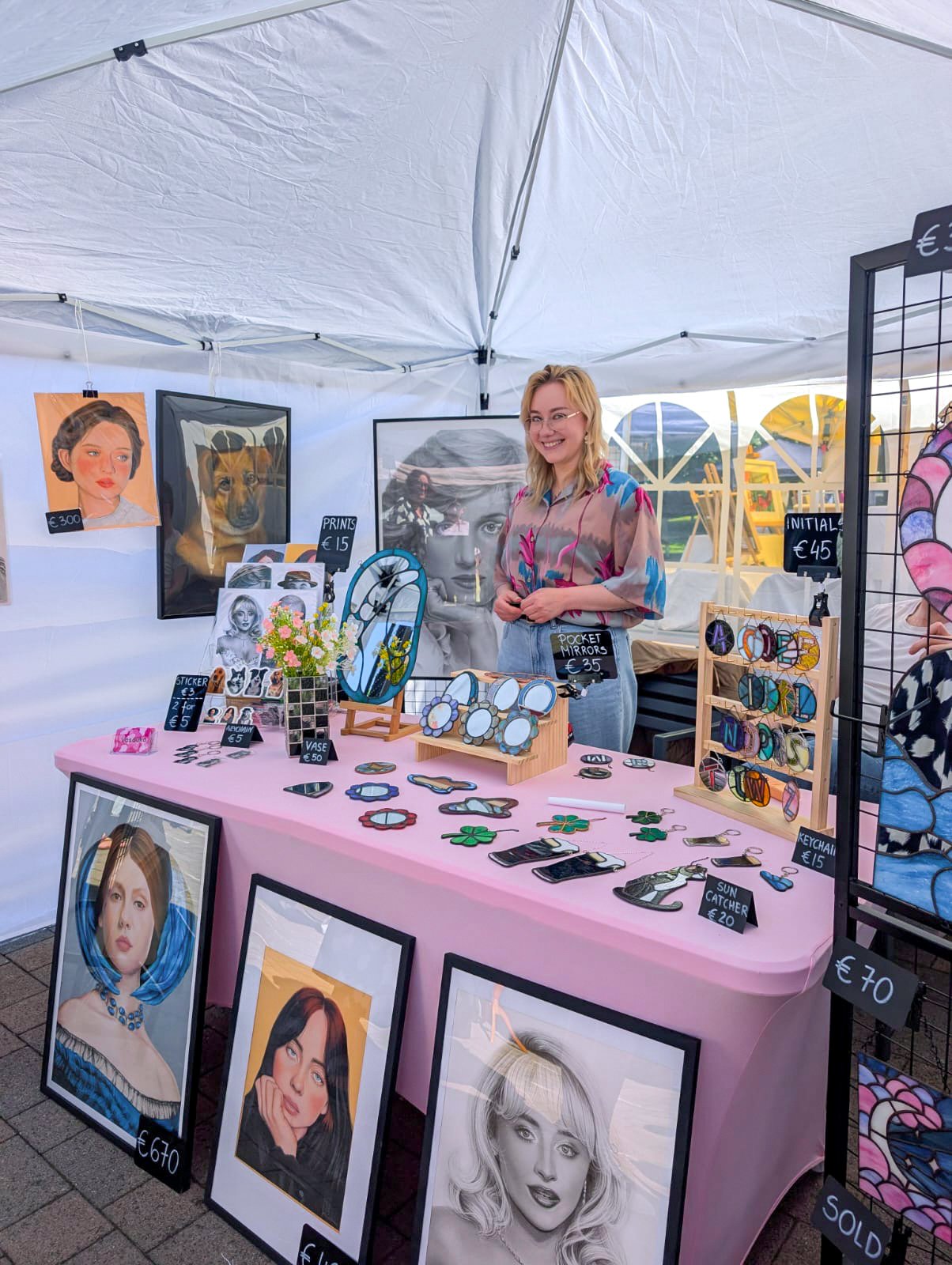 A woman selling artwork and accessories at an outdoor market stall. The stall displays framed portraits, mirrors, jewelry, and accessories on a table covered with a pink cloth.
