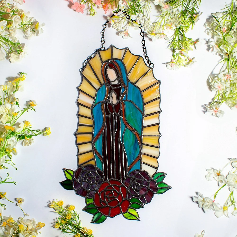 Stained glass depiction of the Virgin Mary surrounded by flowers, hanging among white and yellow blossoms.