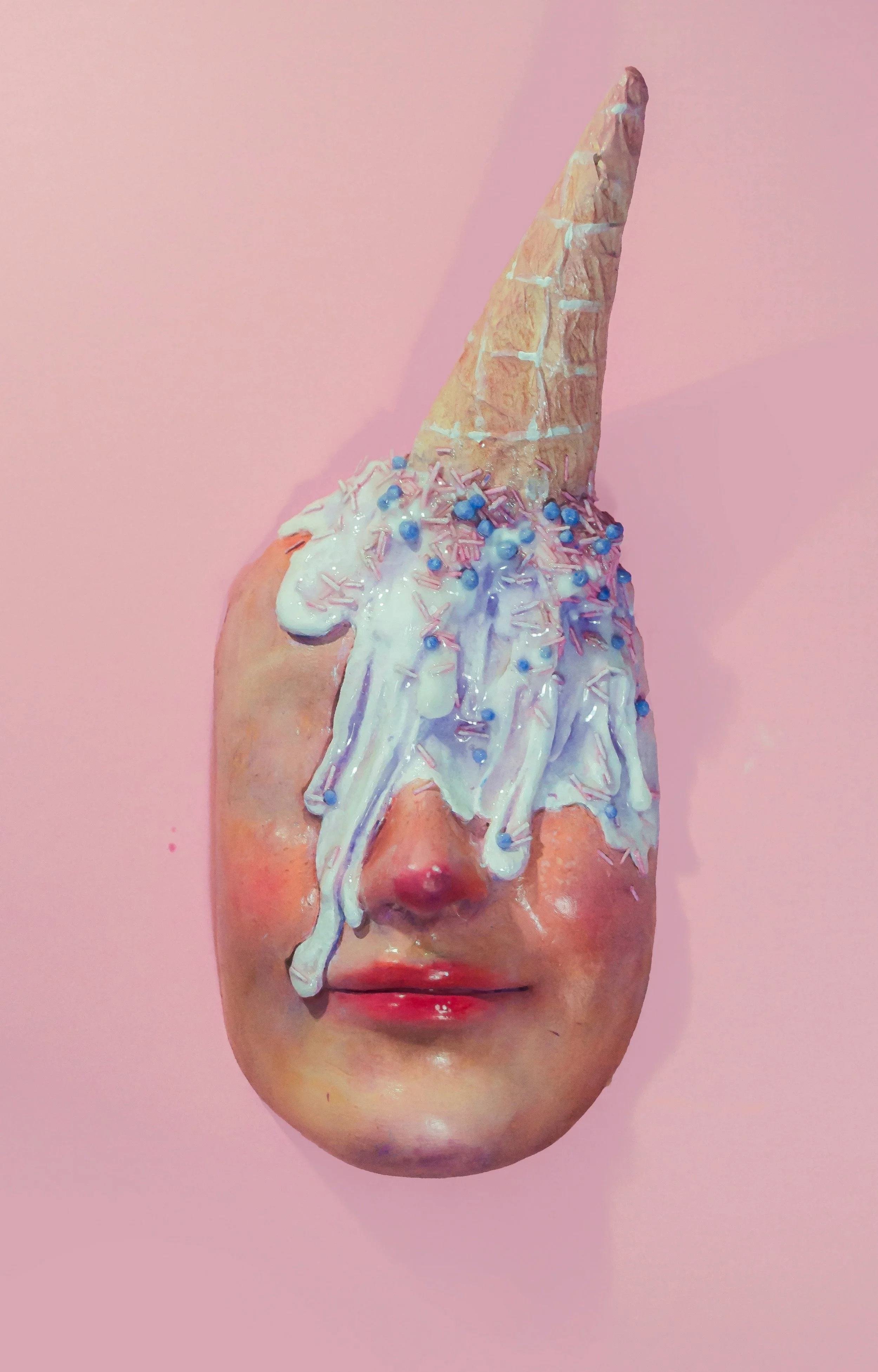 Sculpture of a human face with a splash of melting ice cram with sprinkles on the forehead and eyes on a pink background.