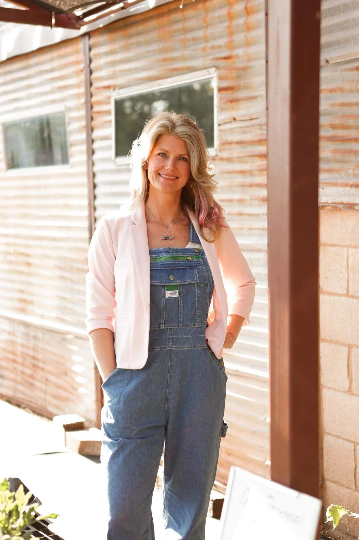 A woman with blonde hair smiling outdoors, wearing a pink blazer and denim overalls, stands near a corrugated metal building.
