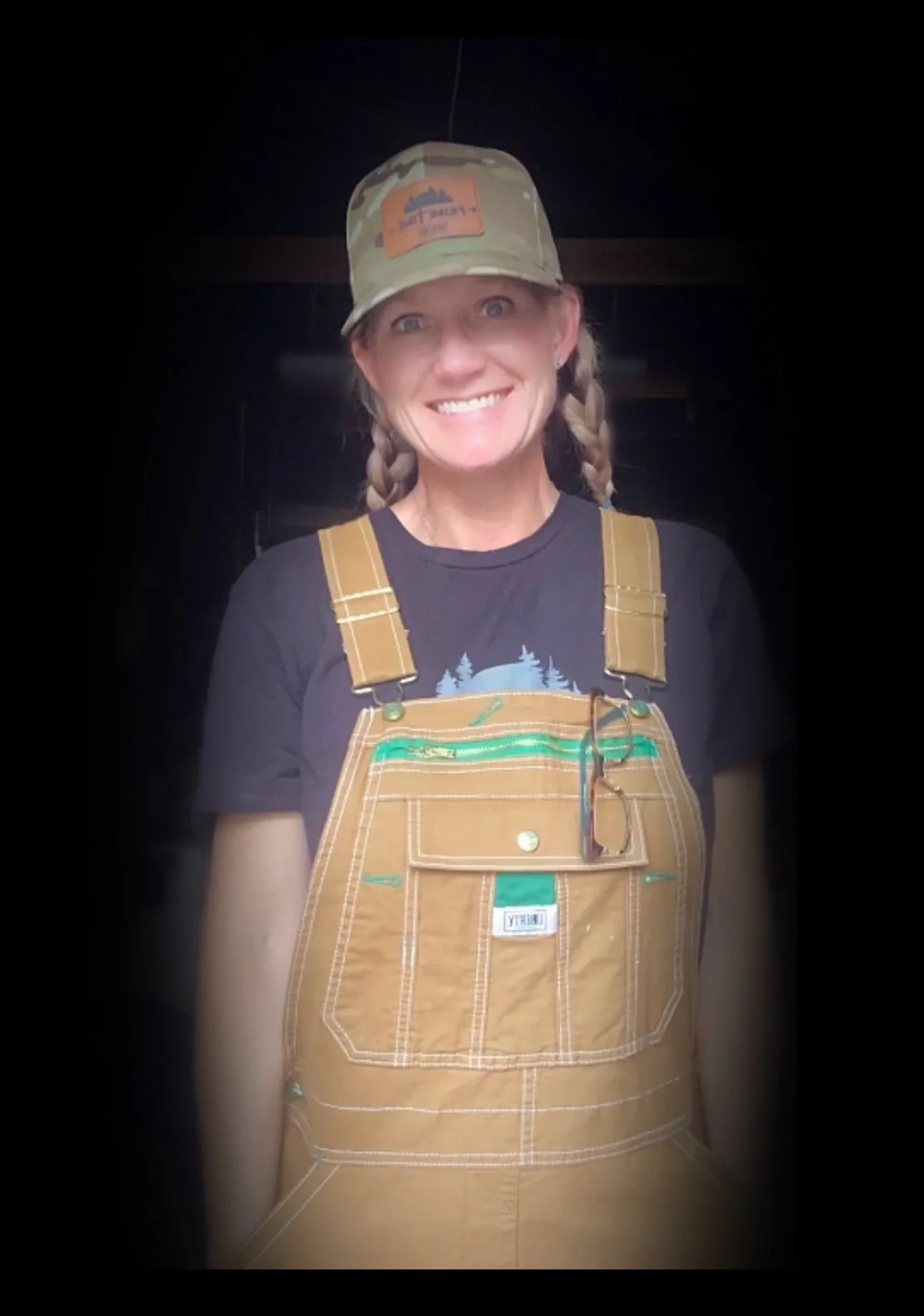 A woman smiling, wearing a camouflage cap, dark t-shirt, and brown overalls with a pair of glasses hanging from the pocket.