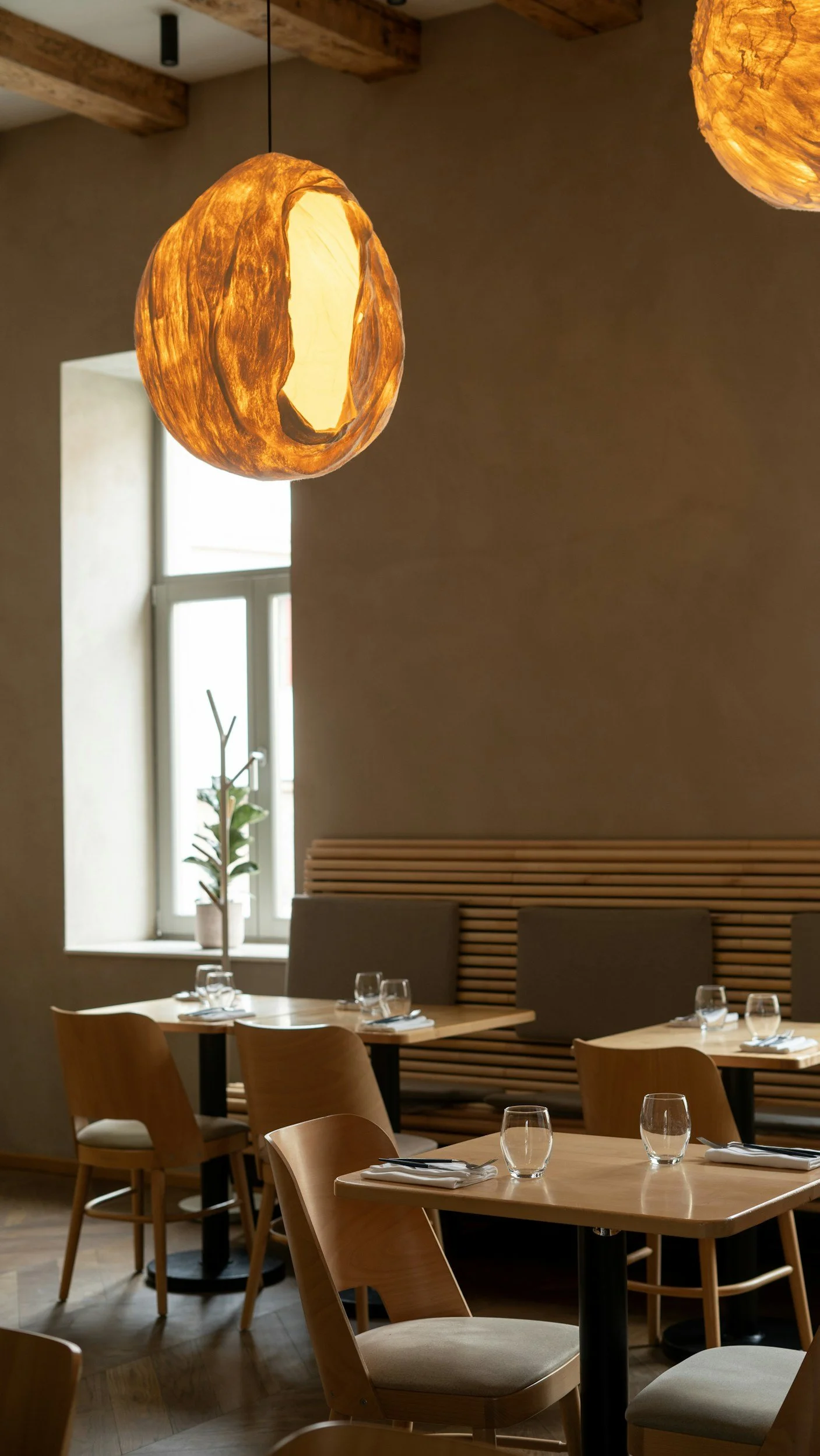 Interior of a modern restaurant with wooden tables and chairs, soft lighting, wall decor, and a window with a potted plant nearby.