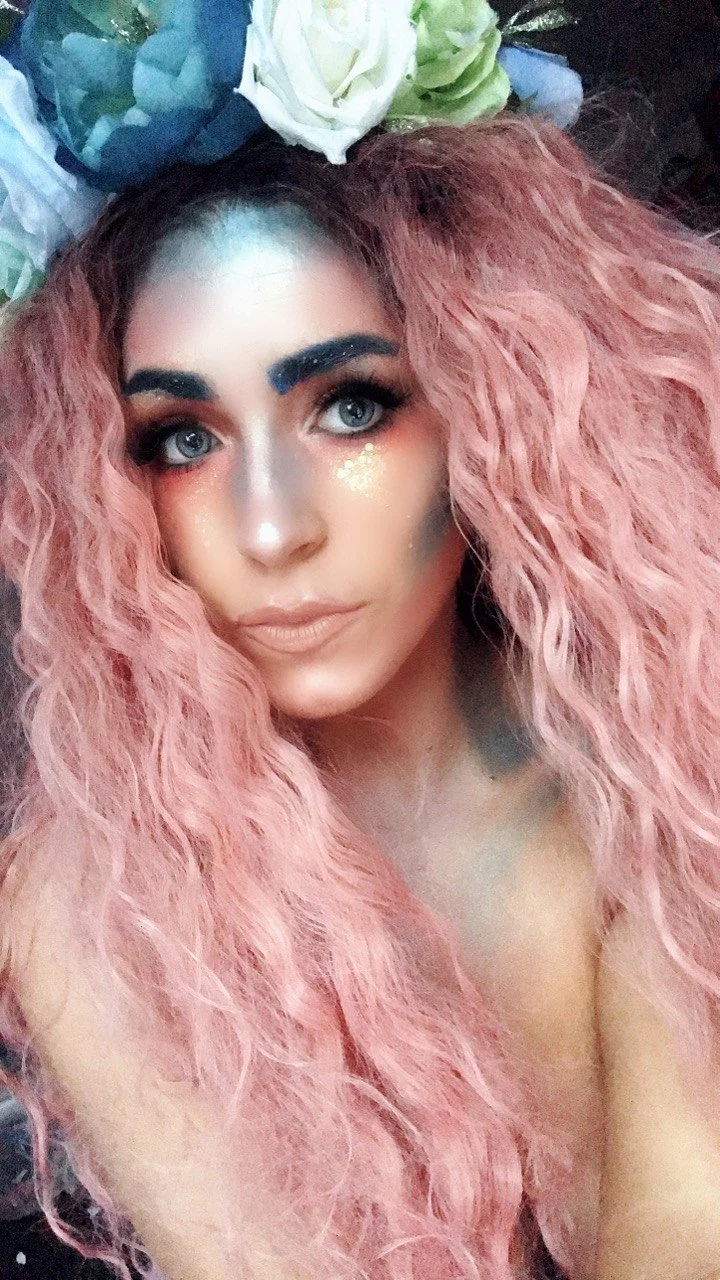 A woman with long, curly pink hair, wearing makeup, and adorned with glitter on her cheek, lying down with flowers in her hair.
