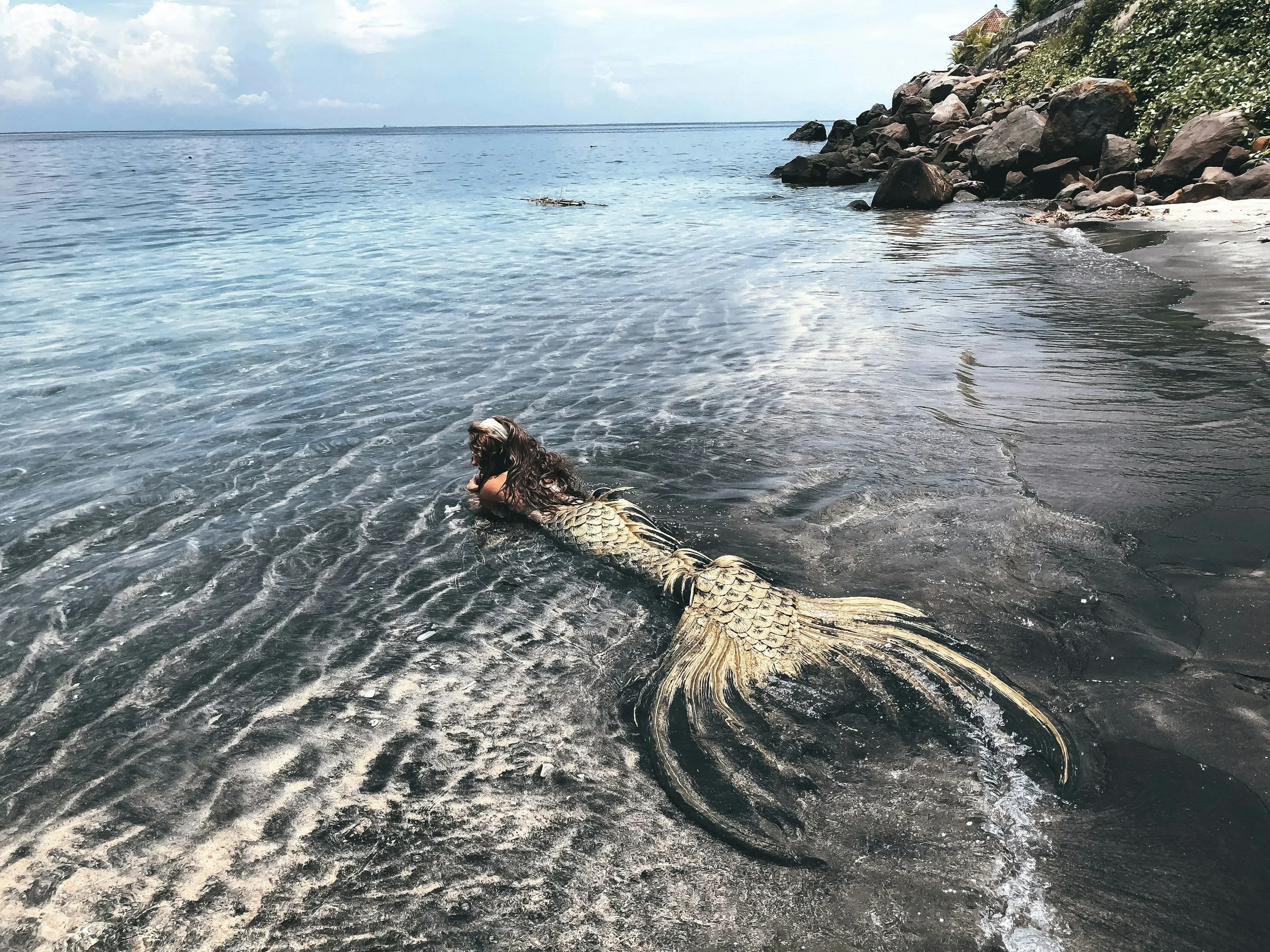 A person with long dark hair dressed as a mermaid in a costume with a fish tail, lying on a shoreline with calm water and rocks in the background. Mermaid Media Social Agency
