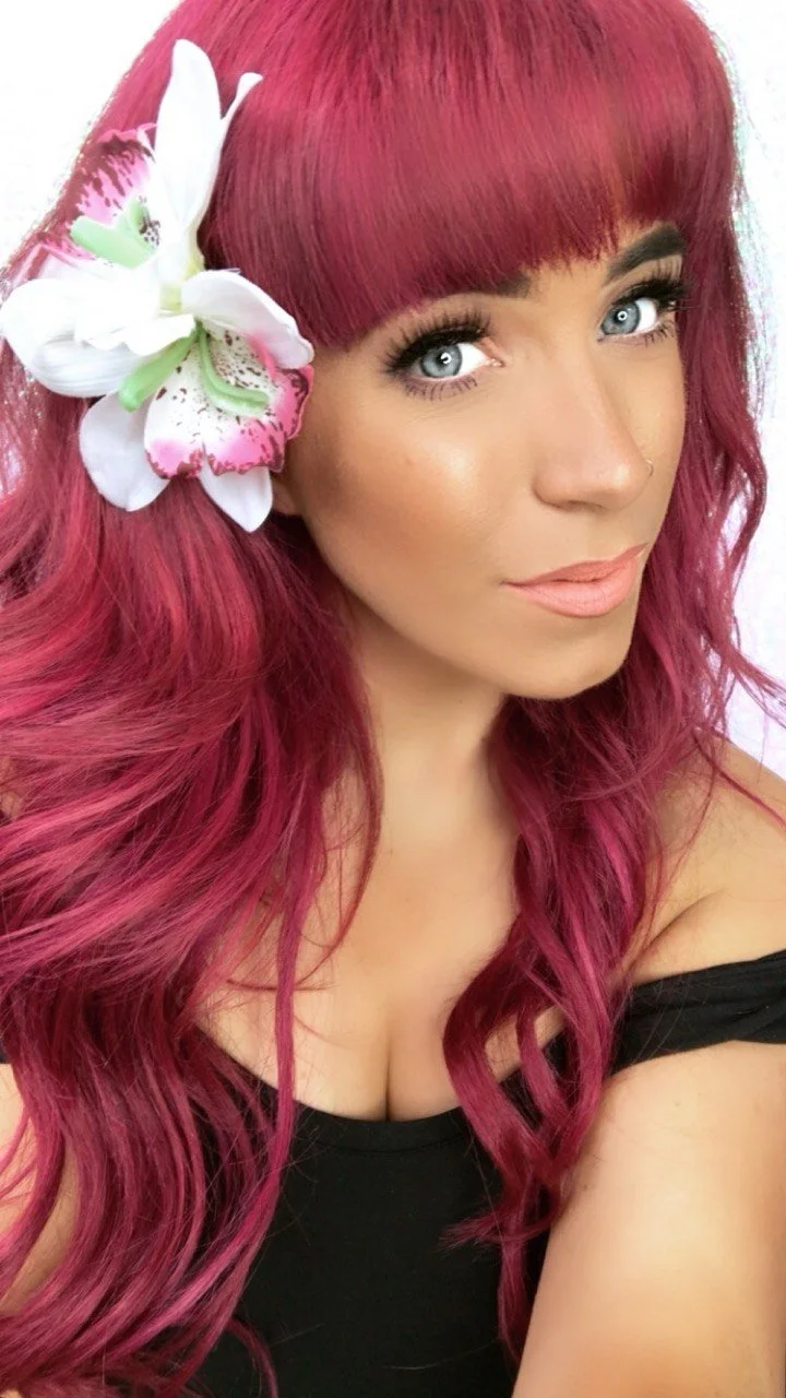 A woman with vibrant pink hair styled in loose waves and straight bangs, wearing makeup with defined eyebrows, long lashes, and peach-colored lipstick, and a large white flower with pink and green accents in her hair, posing in a black off-shoulder top.