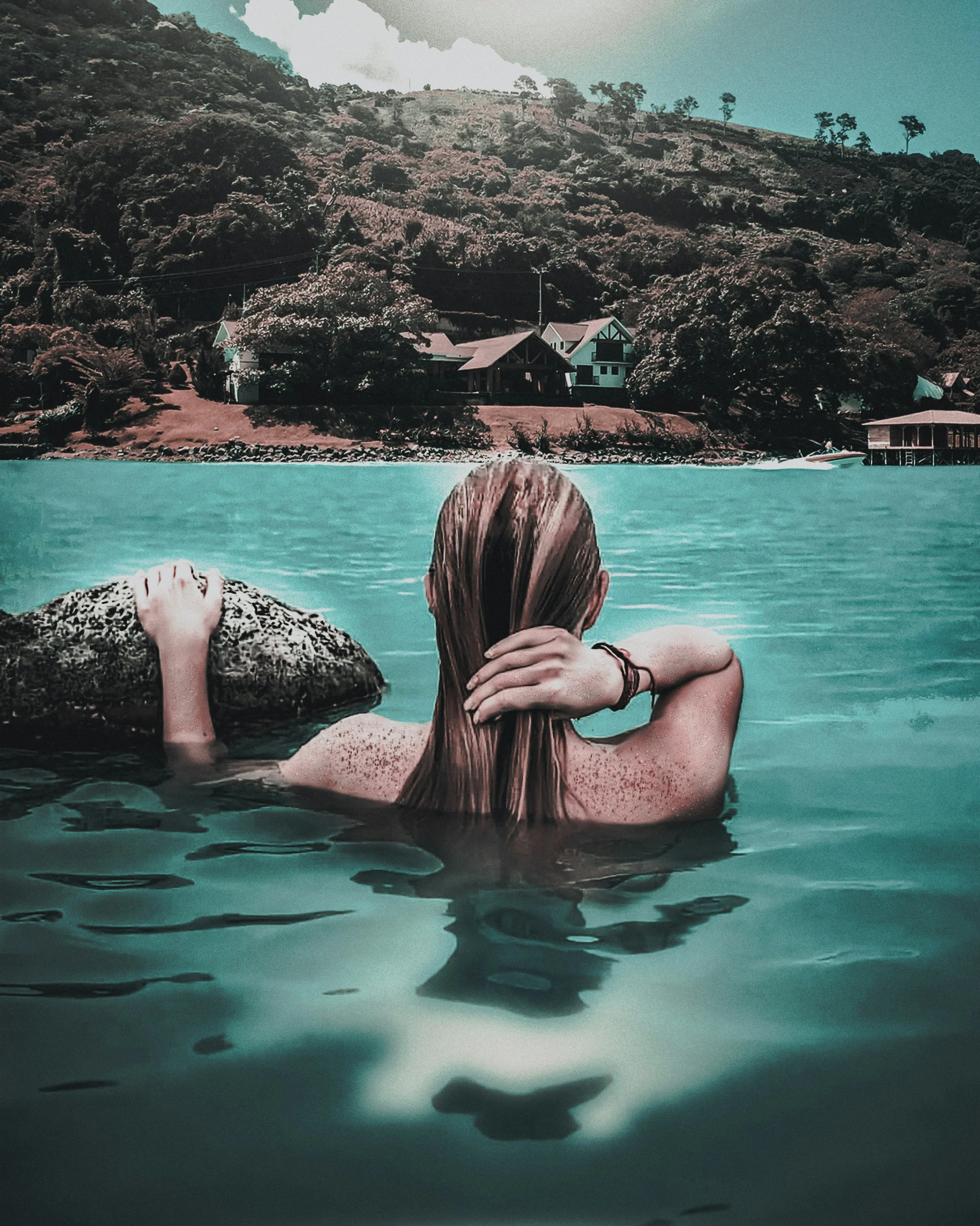 A woman with long, wet, reddish hair is swimming in a body of water, holding a rock with one arm while resting her other arm on her head. In the background, there are houses on a hillside surrounded by trees, and the sky is partly cloudy.