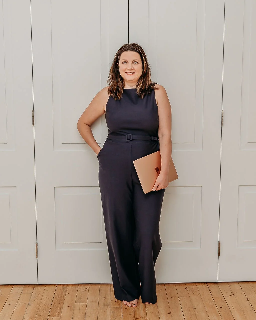 woman wearing a navy one piece, holding a mac computer behind a white wall