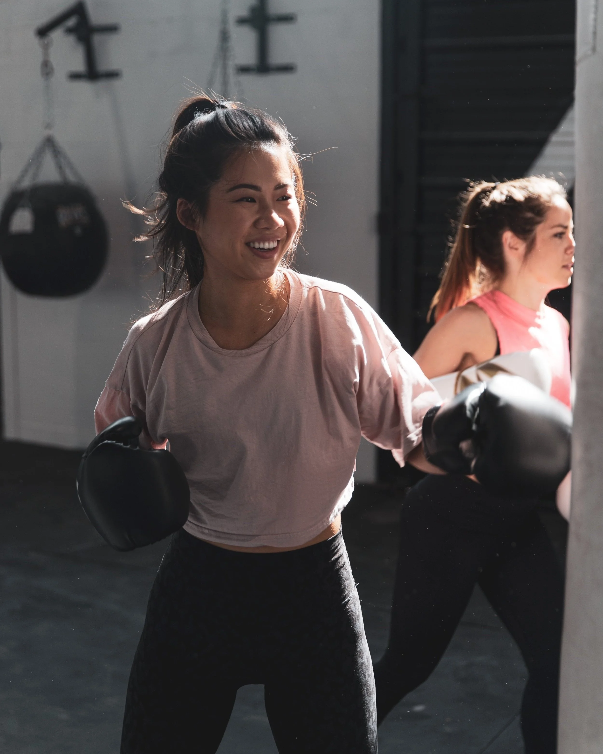 Two women in a gym, one is smiling and wearing a light pink t-shirt with black workout gloves, the other is focused and wearing a pink workout top. Gym equipment is visible in the background.