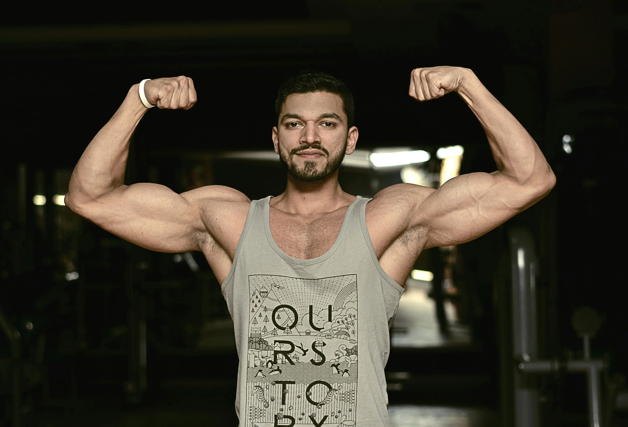A man with a beard and short dark hair flexing his biceps in a gym. He is wearing a light gray tank top with a graphic design. The background is dark with some bright lights.