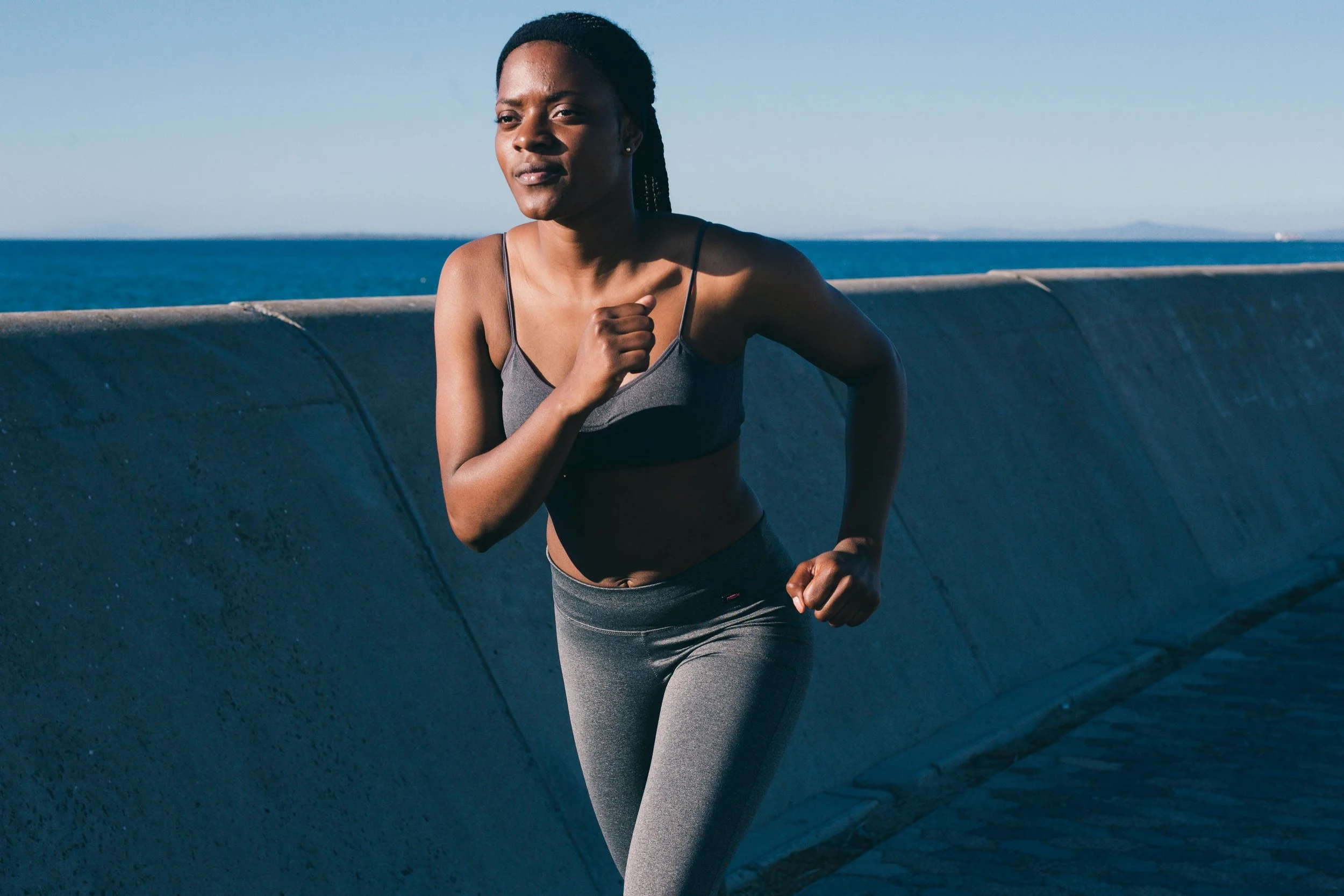 A woman running outdoors near the ocean, wearing a sports bra and gray leggings.
