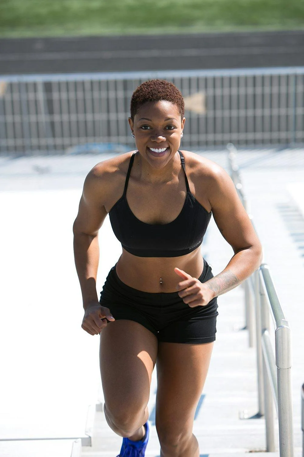 Woman running up outdoor stadium stairs, smiling, wearing black sports bra and shorts, with a belly button piercing.