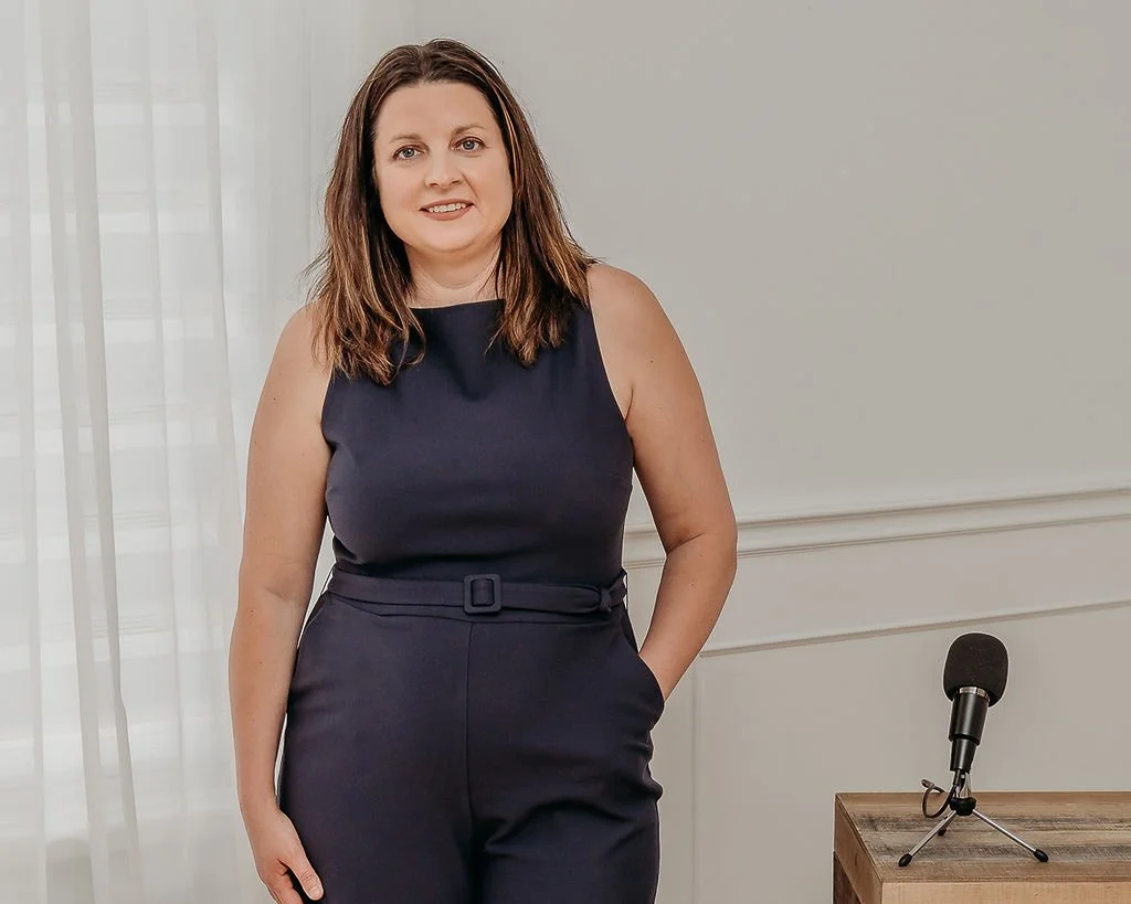 A woman with shoulder-length brown hair wearing a dark sleeveless jumpsuit standing indoors next to a wooden table with a microphone.