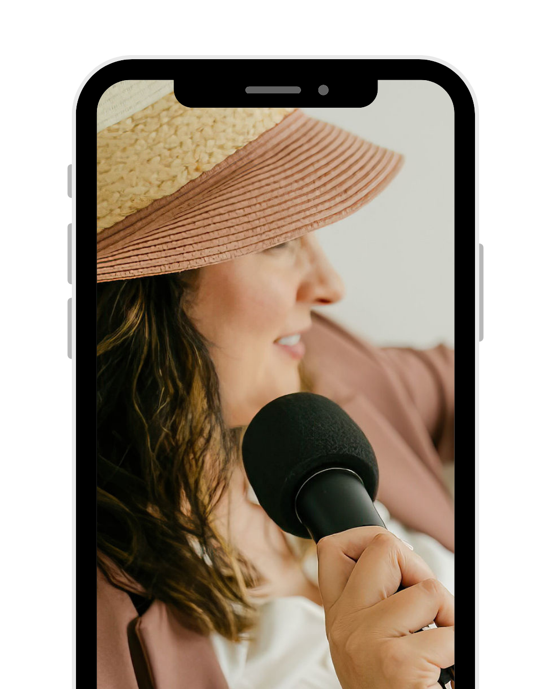 A woman wearing a wide-brimmed hat and a light-colored top, holding a microphone near her mouth, smiling