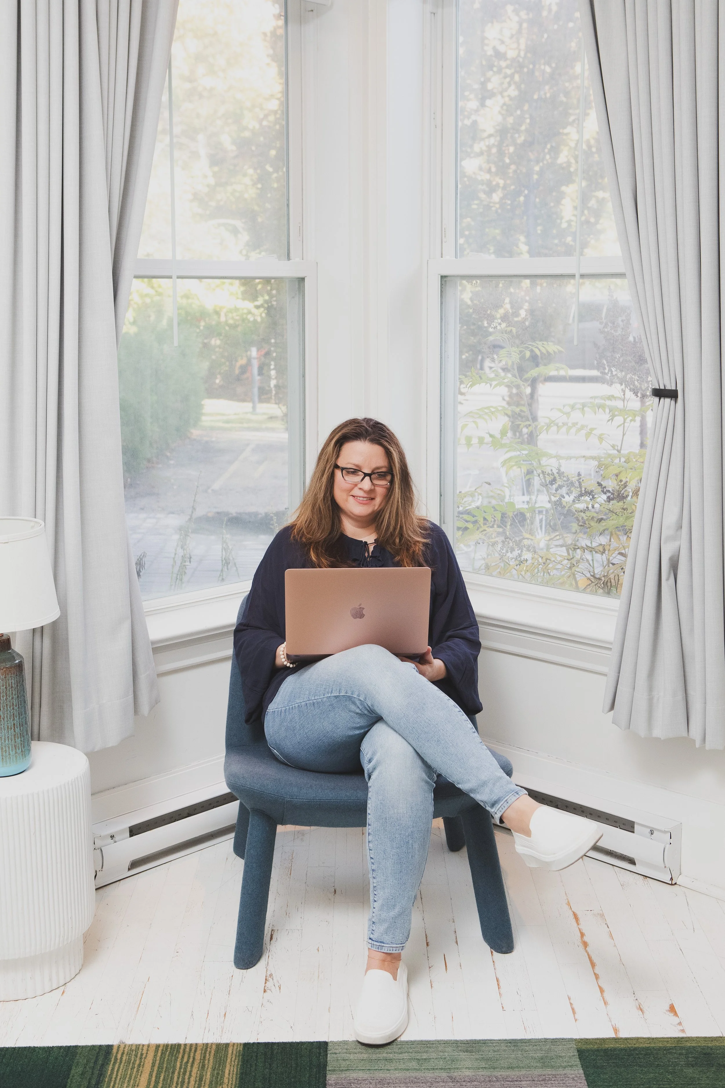 A woman with brown hair, glasses, and a navy blue sweatshirt sitting cross-legged on a blue chair, using a pink laptop. She is in a room with large windows, white curtains, a white side table with a blue vase, and a partially visible white lamp. The floor is white wood, and there is a colorful striped rug at the front of the image.
