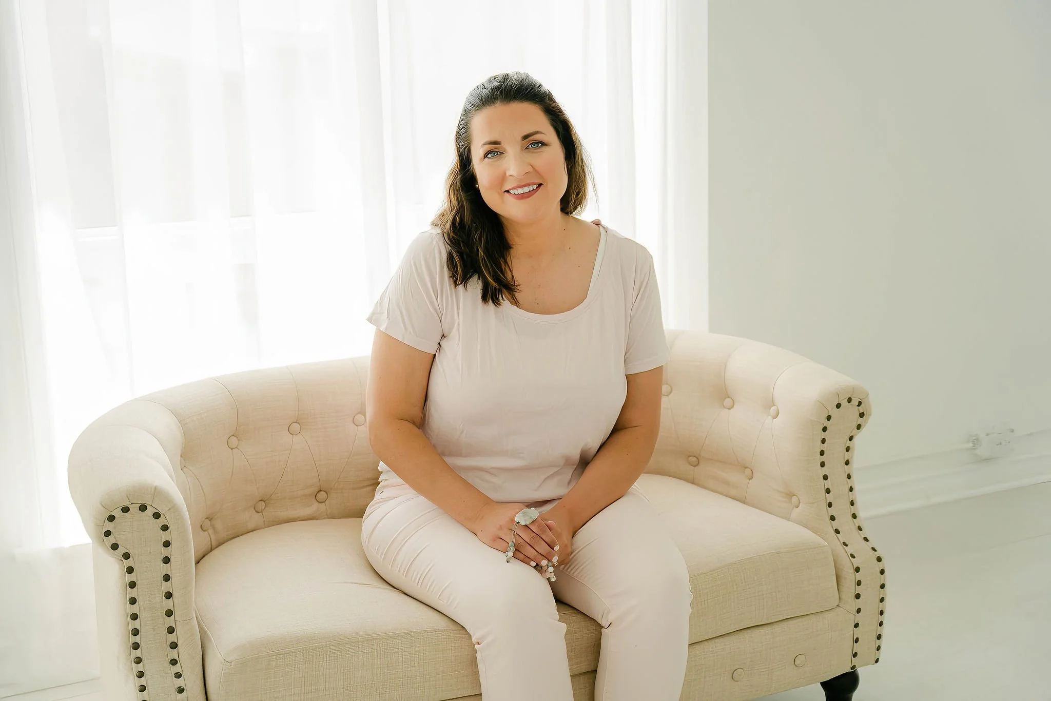 A smiling woman with dark wavy hair, wearing a light-colored top and white pants, sitting on a cream-colored tufted sofa in front of sheer curtains letting in natural light.