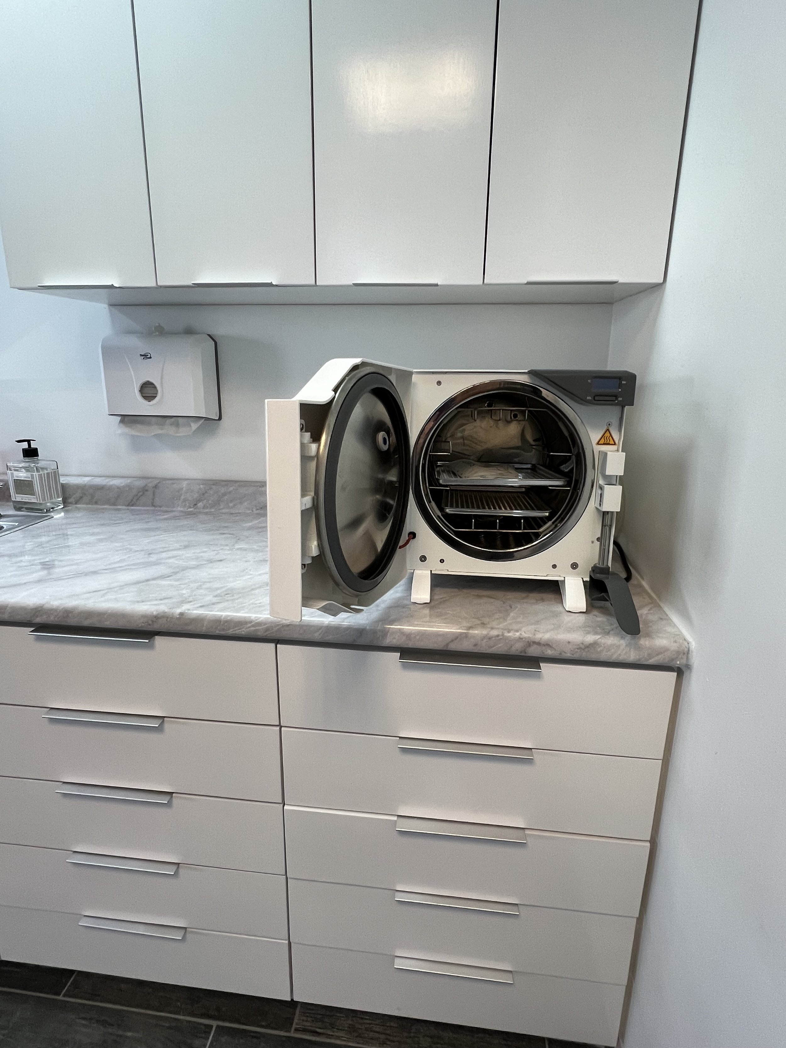 Una máquina de ultrasonido de pie en una encimera de cocina con puertas blancas y un dispensador de jabón en un entorno de clínica o laboratorio.