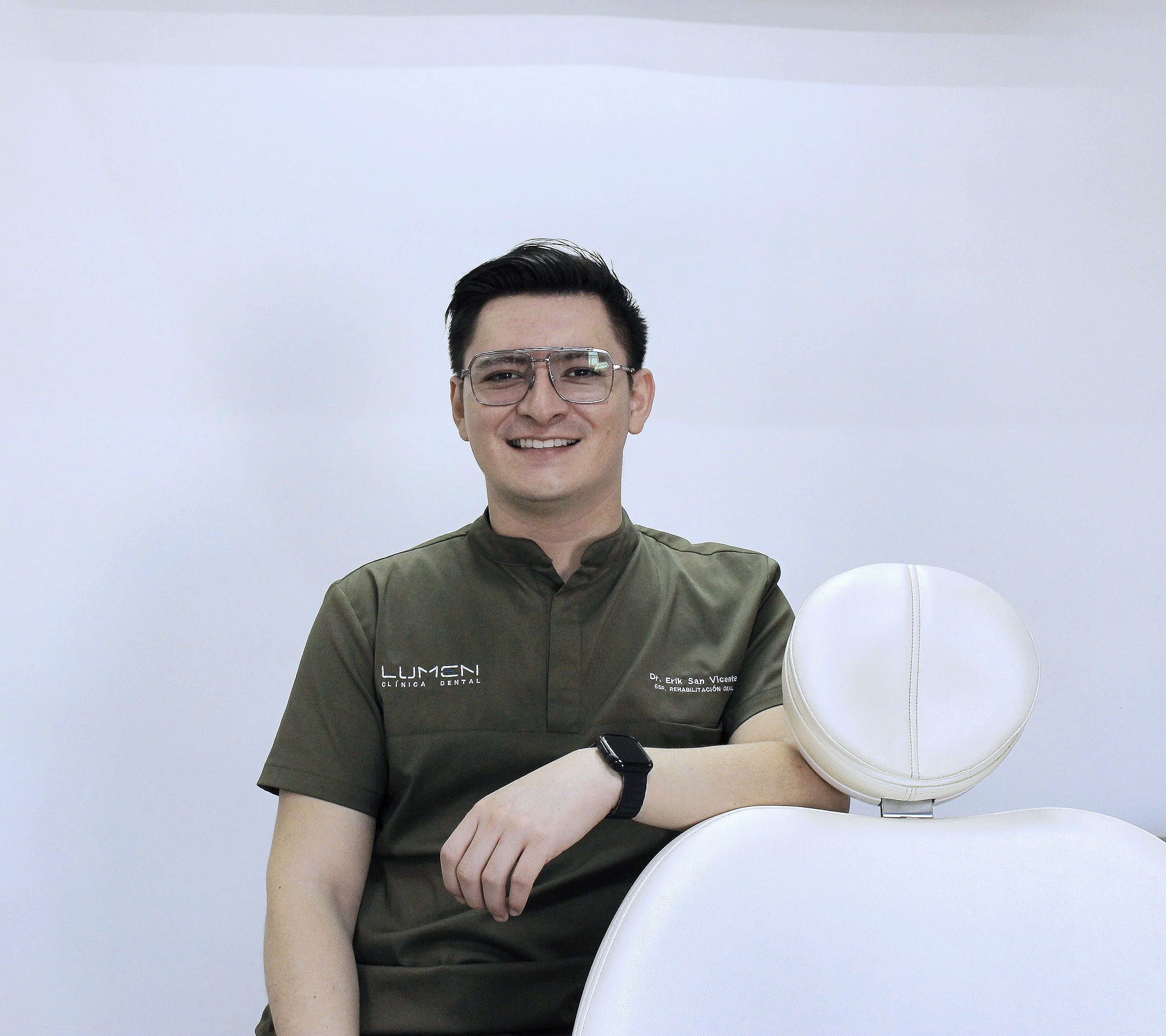 Hombre sonriente con gafas y uniforme en consulta dental, sentado junto a una silla dental blanca.