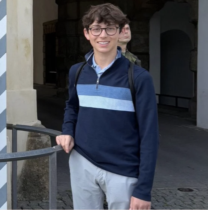 A young man with glasses, curly brown hair, and a smile, standing outdoors in front of a building with archways. He's dressed in a navy blue and light blue quarter-zip sweater, light-colored pants, and carries a backpack.