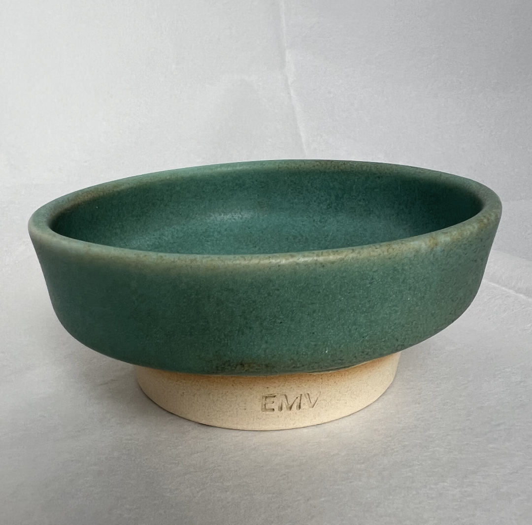 A green ceramic bowl with an unglazed bottom resting on a beige base labeled "EMV" against a plain white background.