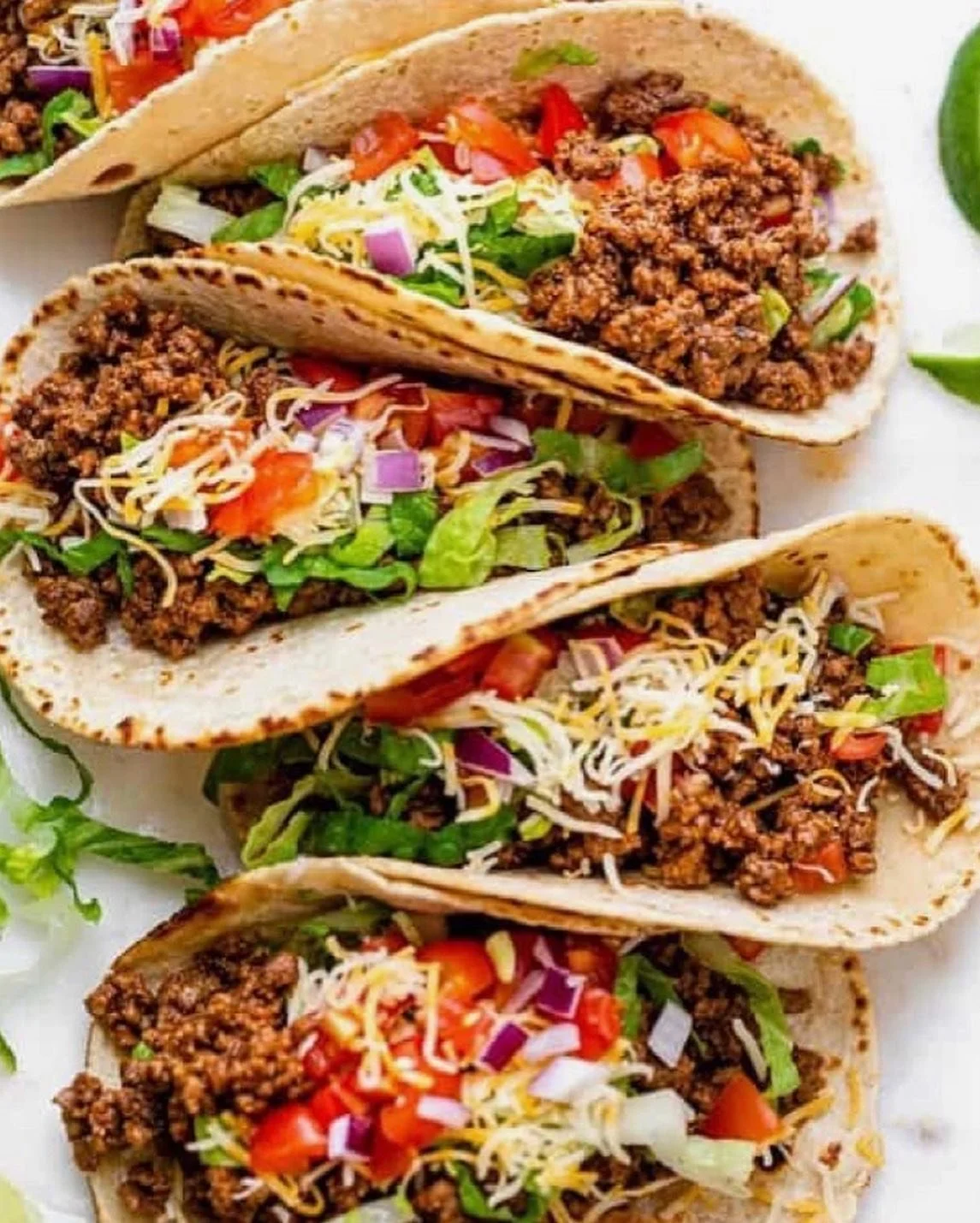 Four tacos filled with seasoned ground beef, shredded cheese, lettuce, tomatoes, and chopped onions in soft corn tortillas.