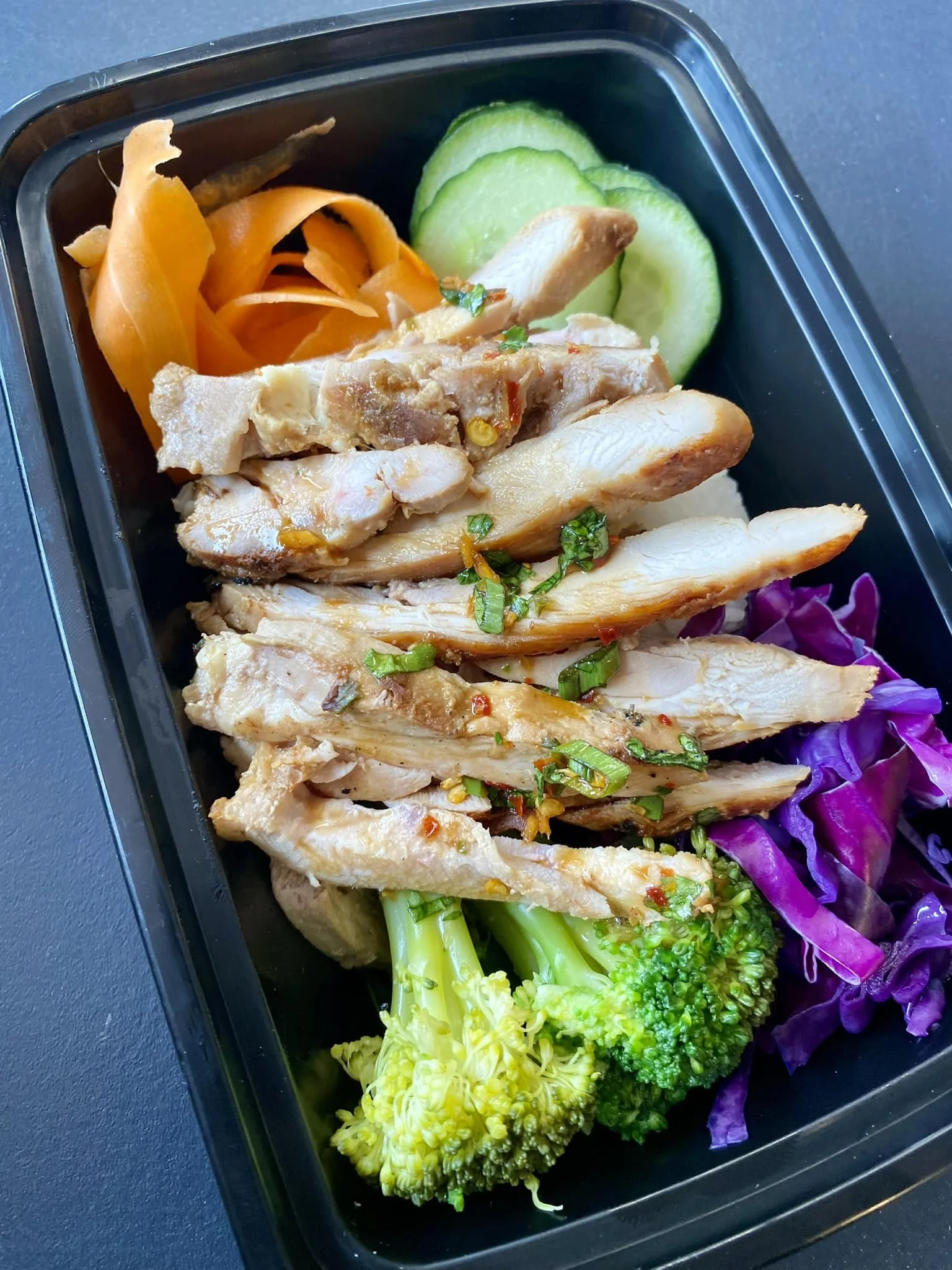 A black meal container holding sliced chicken, broccoli, purple cabbage, cucumber, pickled carrots, and a green bean.