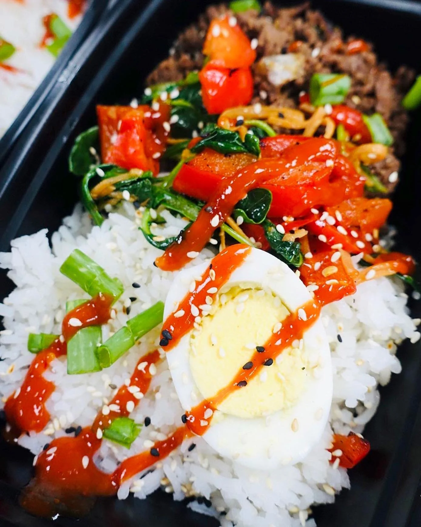 A close-up of a bento box containing white rice, a halved boiled egg, sautéed vegetables, and sliced red peppers, garnished with sesame seeds and drizzled with sauce.
