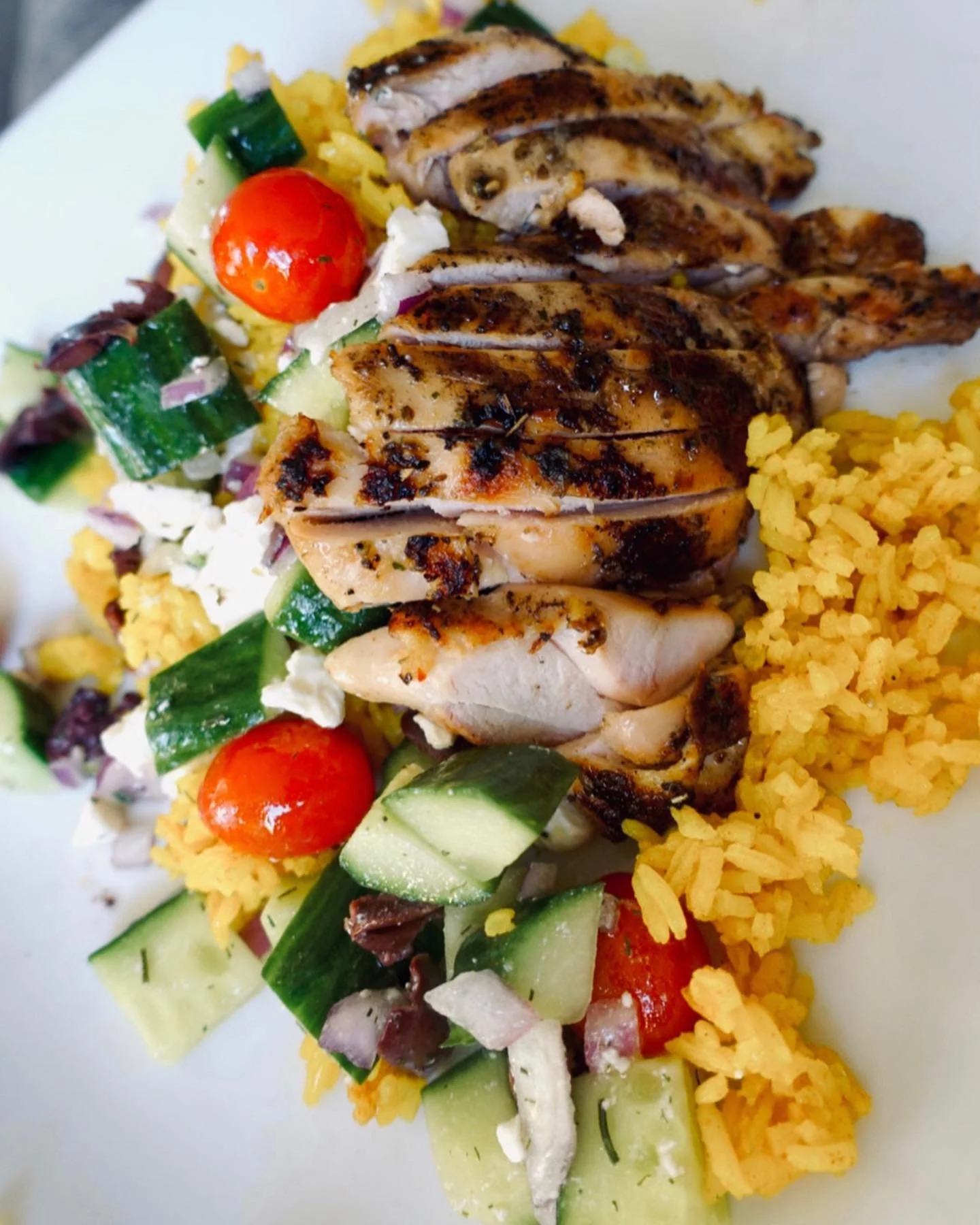Grilled chicken served over yellow rice with cucumber and tomato salad on a white plate.