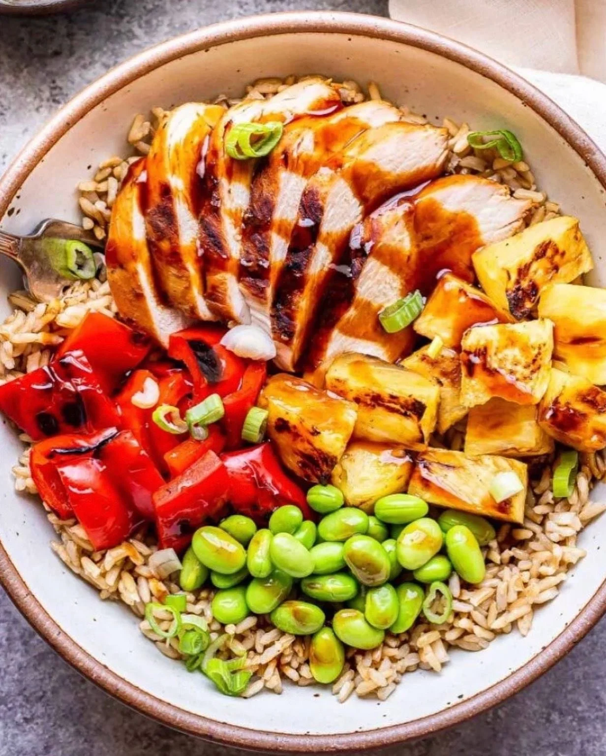 Bowl of cooked brown rice topped with sliced grilled chicken breast, roasted pineapple chunks, chopped red bell peppers, diced green onions, and drizzled with barbecue sauce.