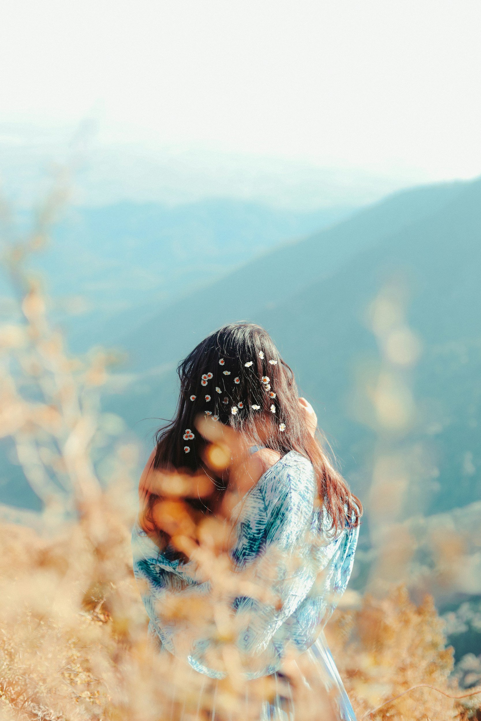 A woman with dark hair and daisies in her hair, standing in a field of tall grass with mountains and a cloudy sky in the background.
