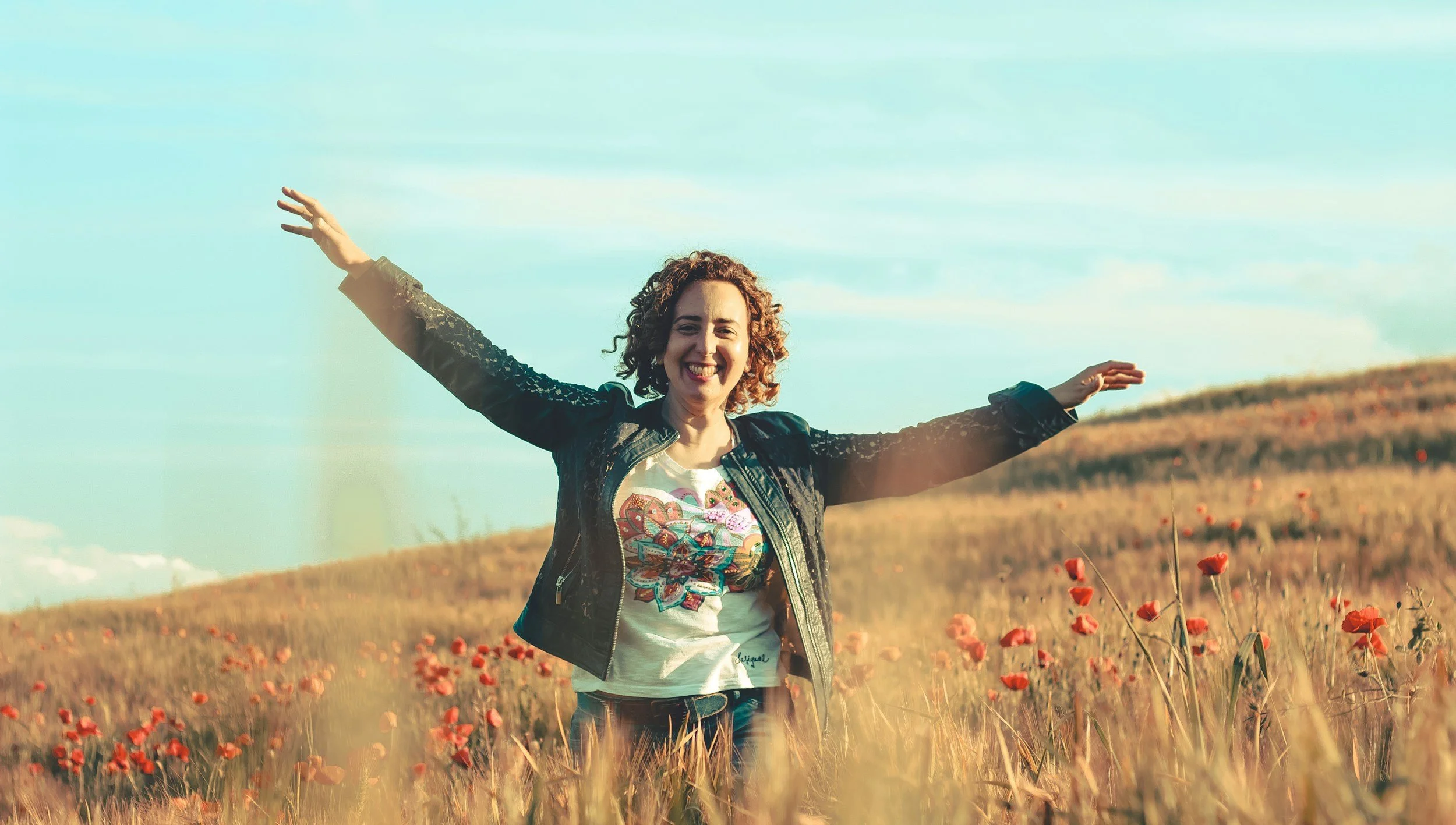 A woman with curly hair smiling and extending her arms outward in a field of orange poppies during daytime with a blue sky and some clouds in the background.