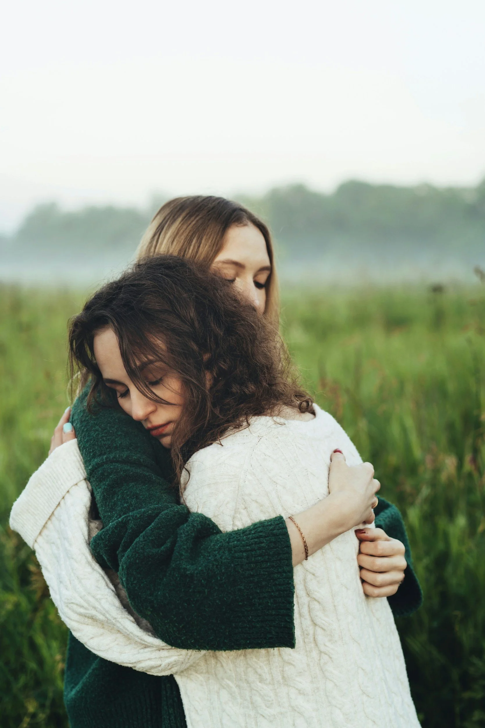 Two women embracing outdoors, with one woman's face resting on the other's shoulder, surrounded by green fields and trees.