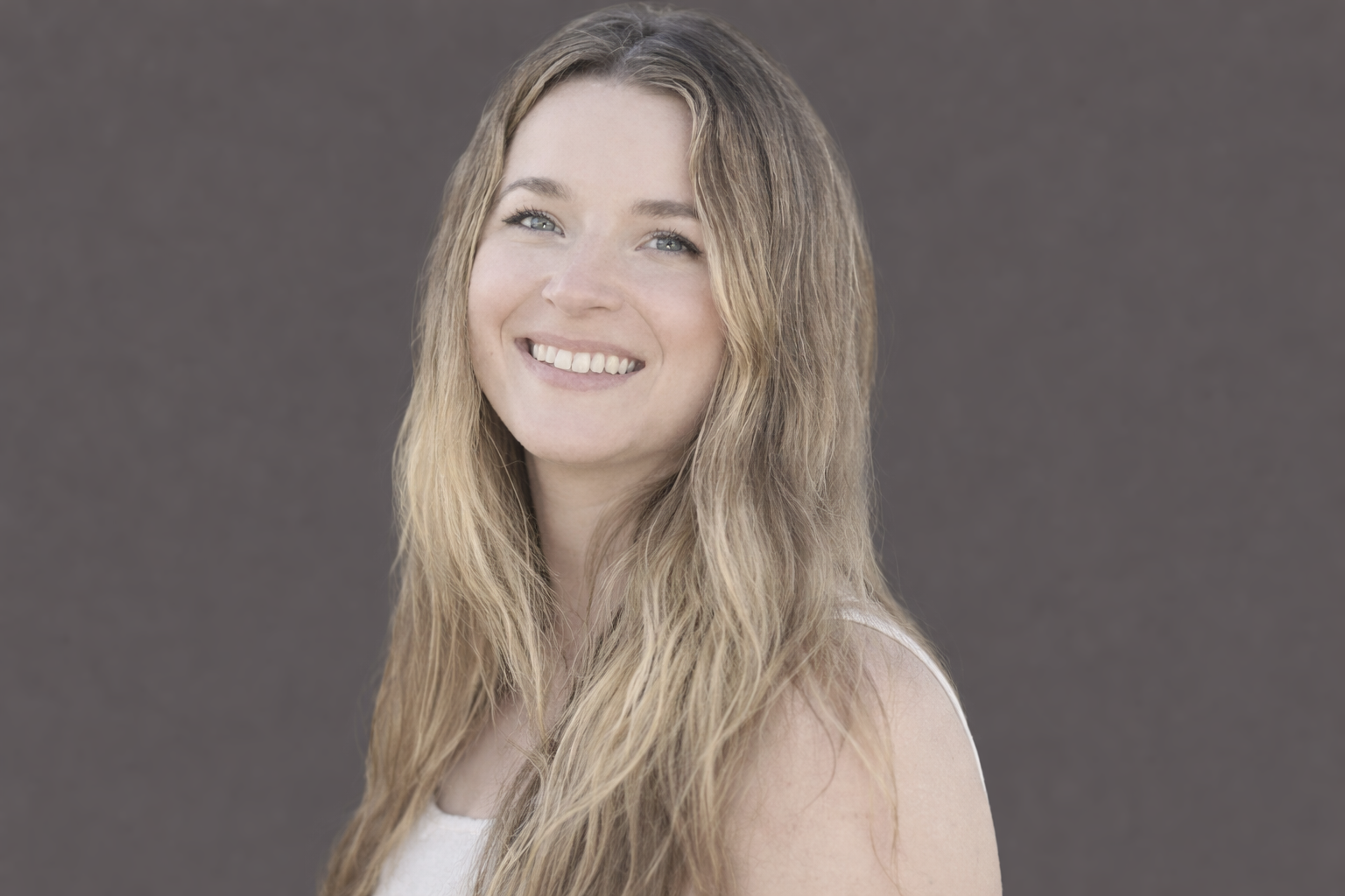 A portrait of a smiling woman with long, wavy blonde hair, blue eyes, and fair skin, wearing a white top against a gray background.