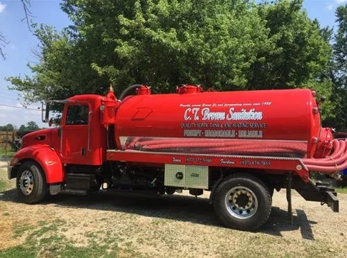 Red septic truck with multiple hoses, parked outdoors on dirt and grass, with green trees and blue sky in the background.