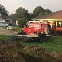 A flatbed trailer parked on a grassy area next to a house, with a red fire truck and other vehicles in the background.
