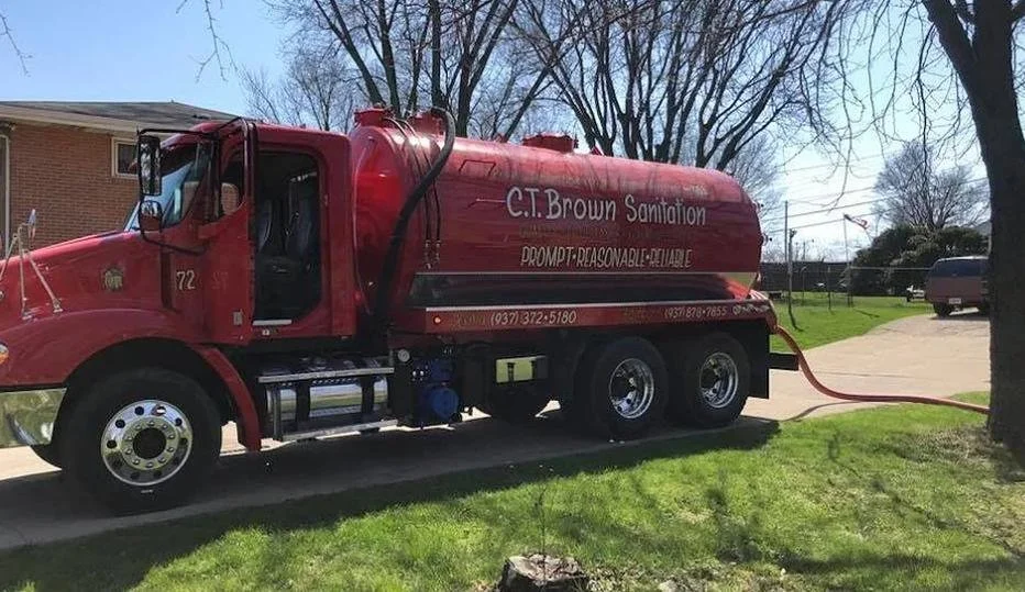 Red sanitation truck parked on grass with a hose attached, in front of a brick building and leafless trees.