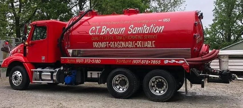 Red septic tank truck with white lettering advertising C.T. Brown Sanitation, displaying phone numbers, and slogan 'Prompt, Reasonable, Reliable' parked on a gravel surface with trees and a building in the background.
