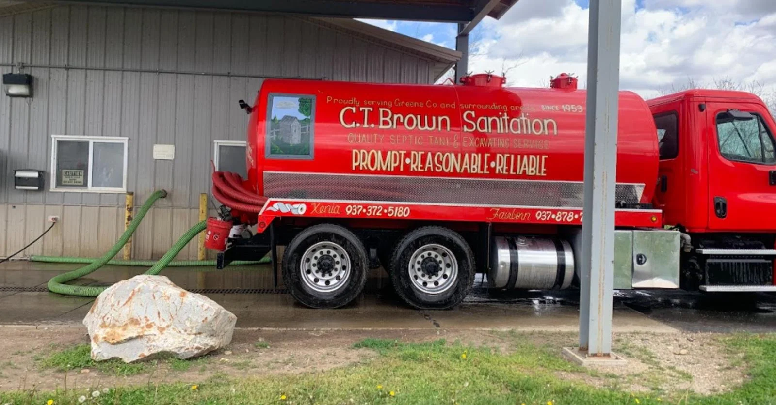Red septic tank and excavation service truck parked outside a building, with hoses connected, and a large rock on the grass nearby.
