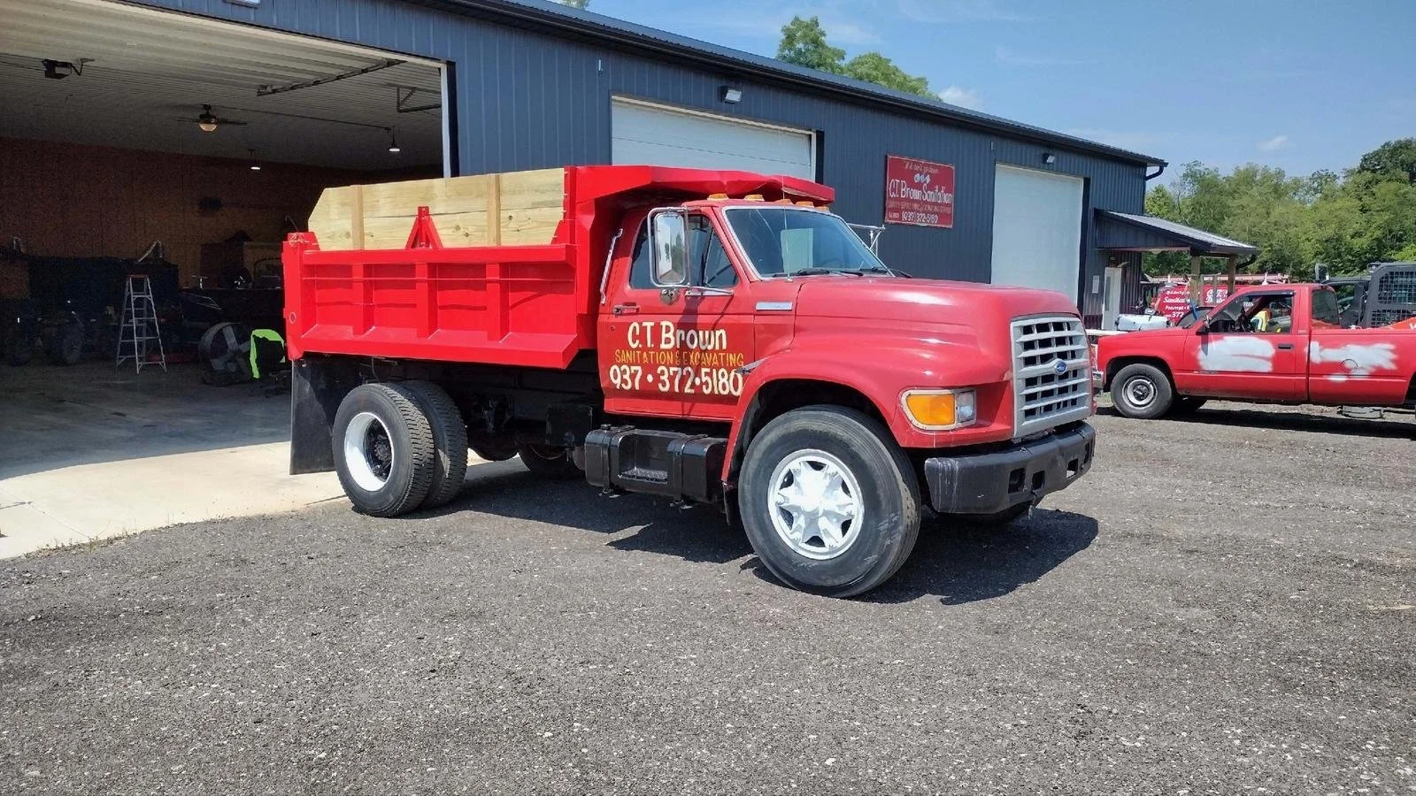 Red dump truck parked outside a garage with a partially open door, showing an interior with several vehicles and a person working. The truck has a company logo and contact information on the side. Another red pickup truck with white paint patches is 