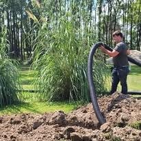 A man working at a construction site near a pond, holding a large flexible pipe or hose, with tall grass and trees in the background.