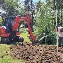 A person working near a small orange excavator in a garden or yard, digging into the soil amidst trees and greenery.